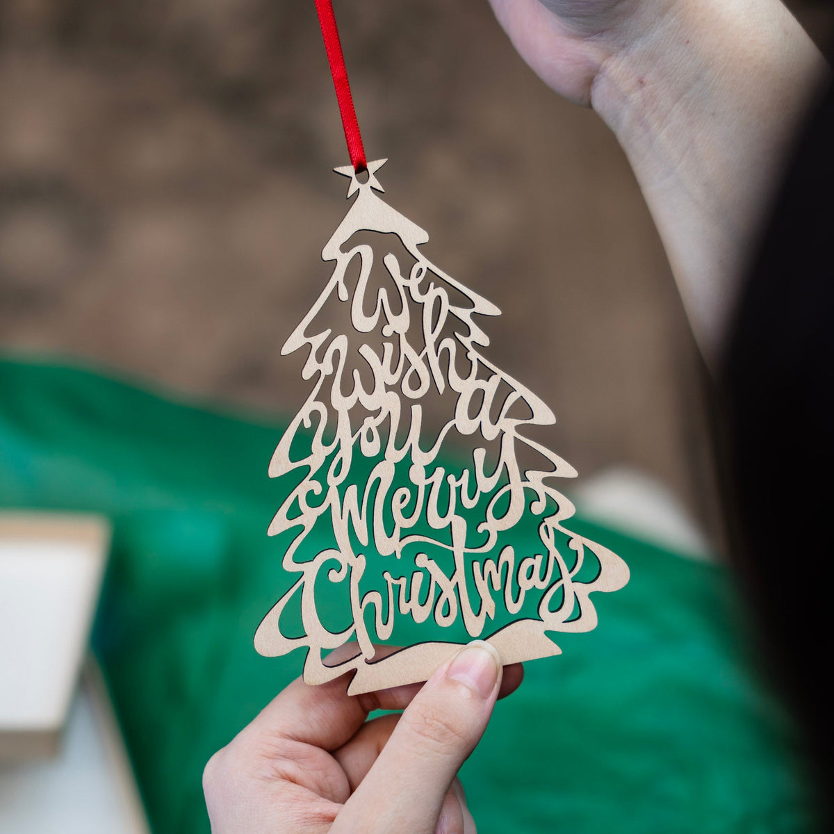 A close-up of a hand holding a laser-cut wooden Christmas tree ornament with 'We Wish you a Merry Christmas' written in the branches, suspended from a red ribbon. A person's other hand is holding it up against a soft-focus background with green tissue paper and gift box.