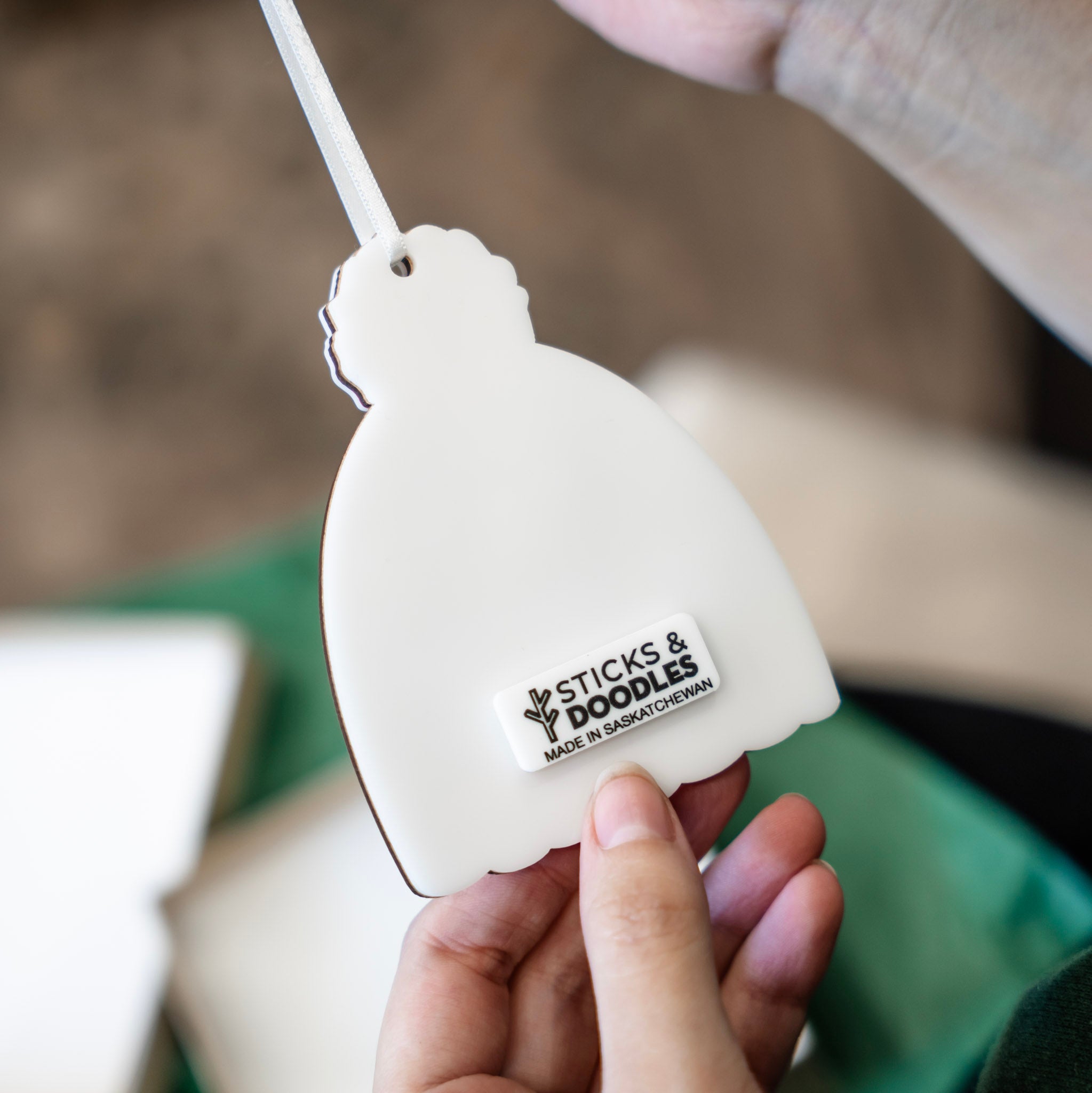 A hand holding the back of a white, toque shaped ornament with a tag that has the words 'STICKS & DOODLES' on it set against a softly blurred background.