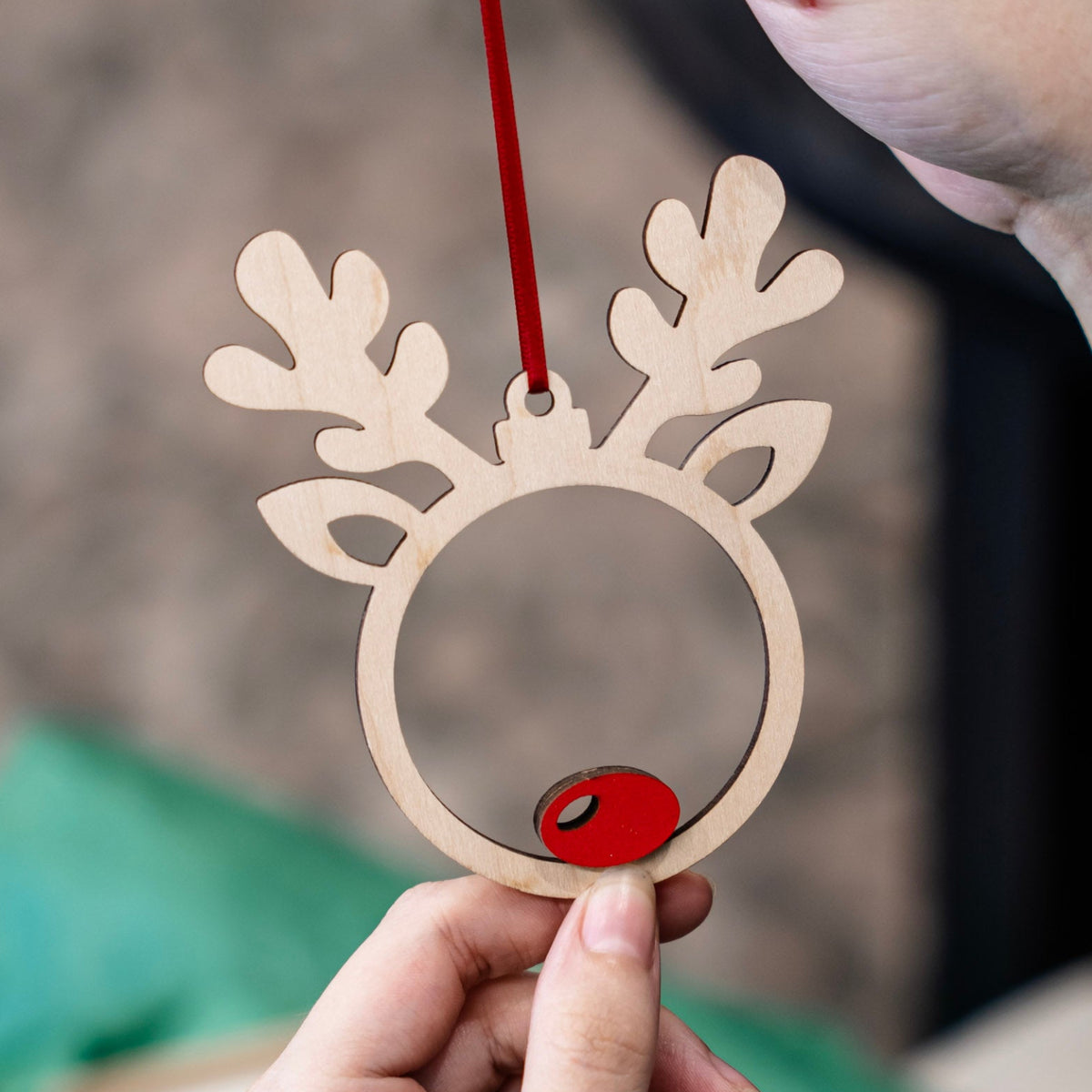 A wooden reindeer ornament held by hands, featuring a natural light wood, red hanging ribbon, and a red rudolph nose.