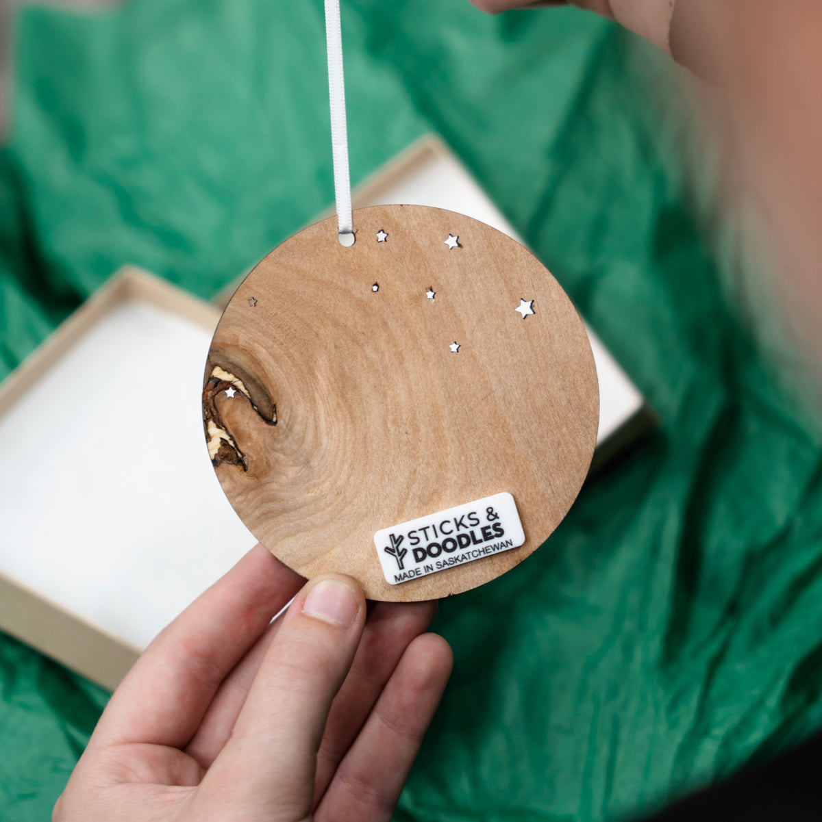 A person holds the back of a wooden hanging ornament with star cutouts and a visible 'STICKS & DOODLES' label, against a green crinkled fabric background with part of a box visible.