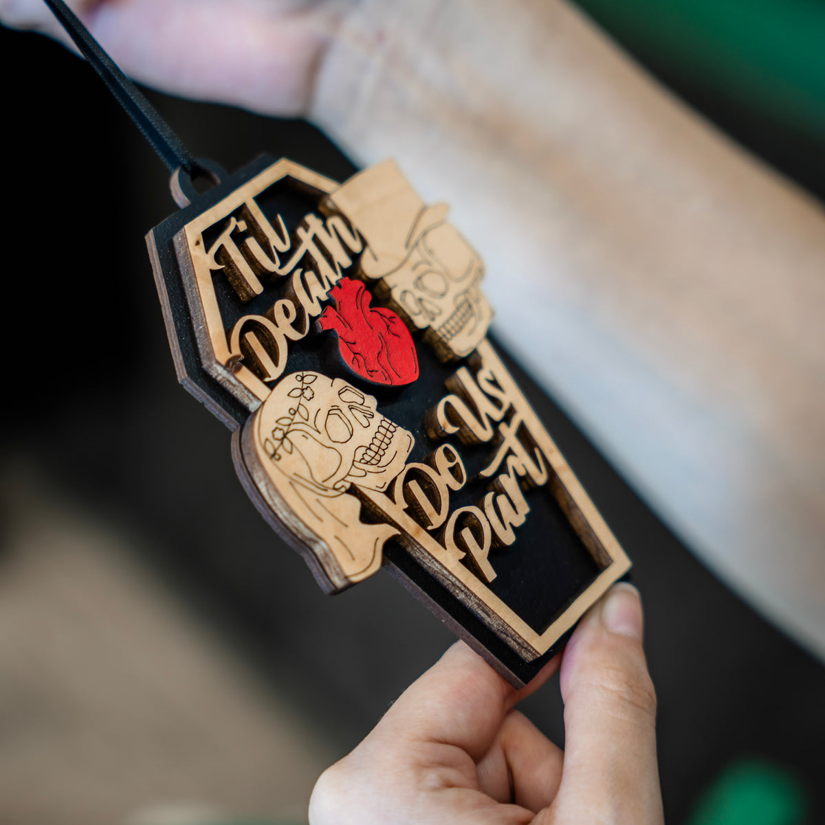 A close up of a coffin-shaped wooden ornament with the phrase 'Til Death Do Us Part' in laser-cut wood, featuring a red anatomical heart and skull bride and groom details, against a blurred background.