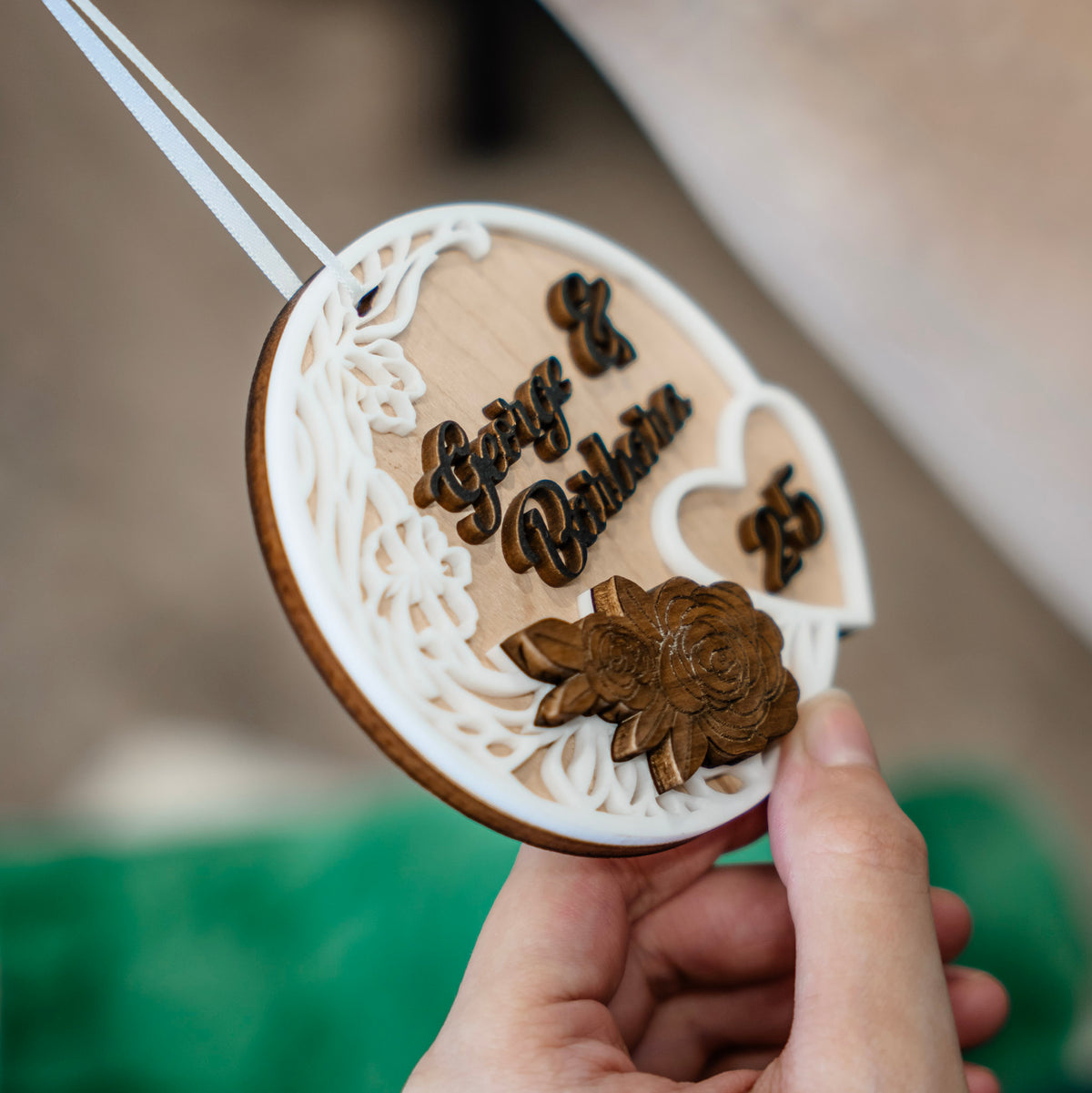 A close-up of a hand holding a personalized wooden ornament. The round, laser-cut wooden ornament features white acrylic floral cutouts, laser cut black text reading 'George & Barbara', with the number 25 in a small heart, and a 3D wooden rose.