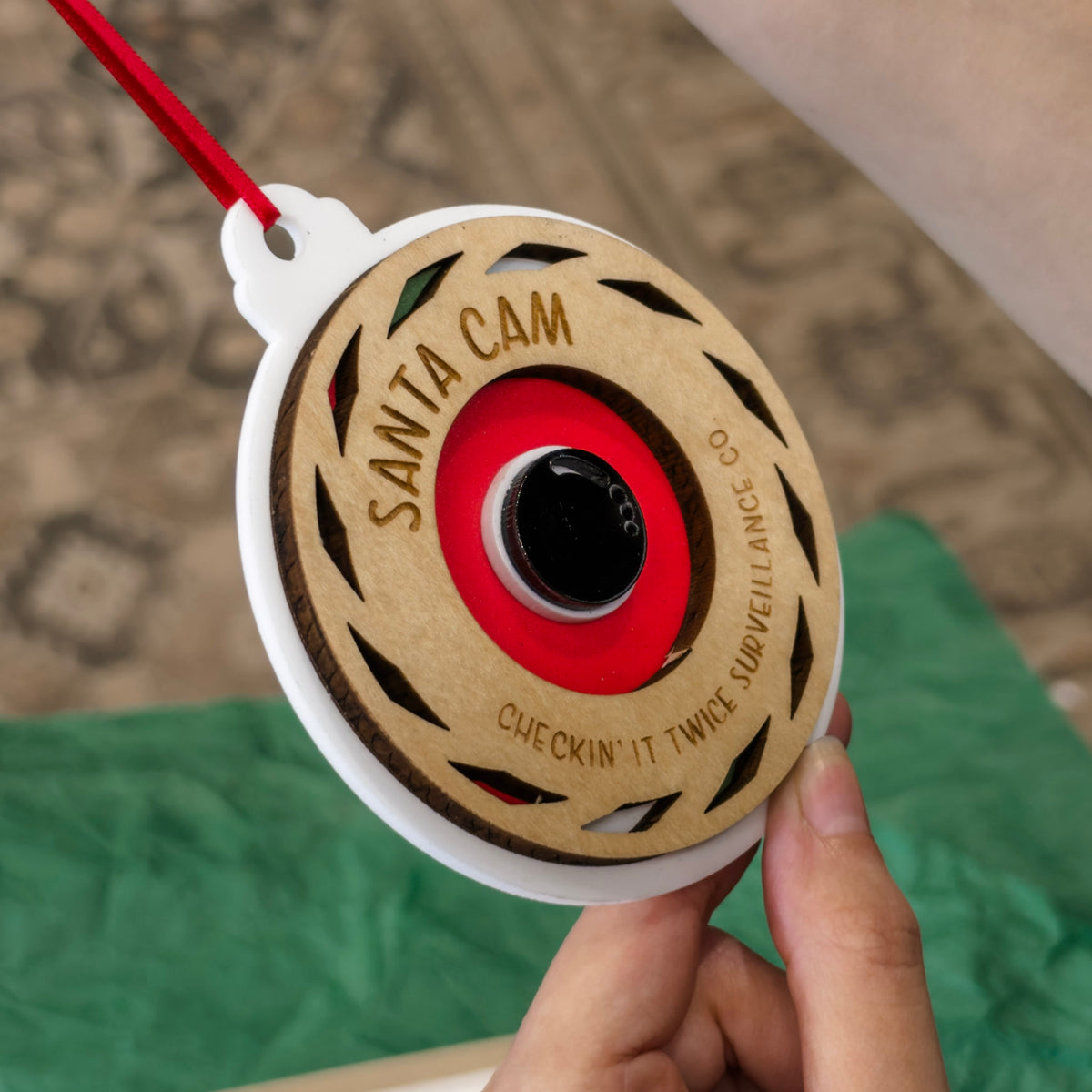 A hand holding a round wooden Santa Cam ornament with a red hanging ribbon. The ornament features a natural wood face with a red and black camera lens design in the center, surrounded by the words 'SANTA CAM' and 'CHECKIN' IT TWICE SURVEILLANCE CO.