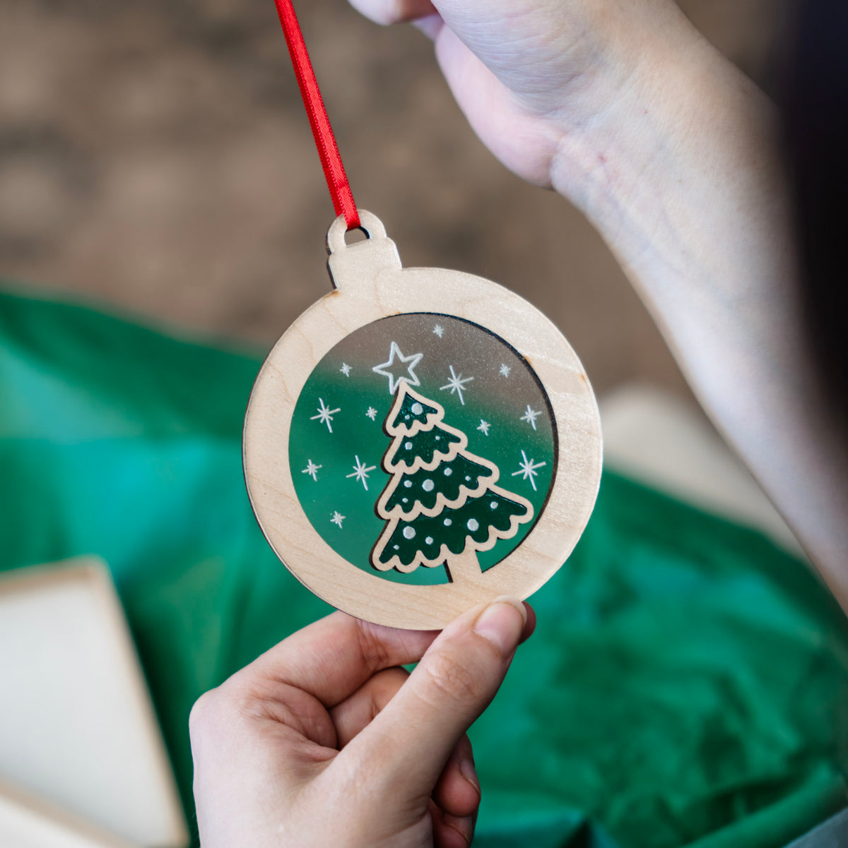 A person holds the back of a round wooden and acrylic Christmas  tree ornament .