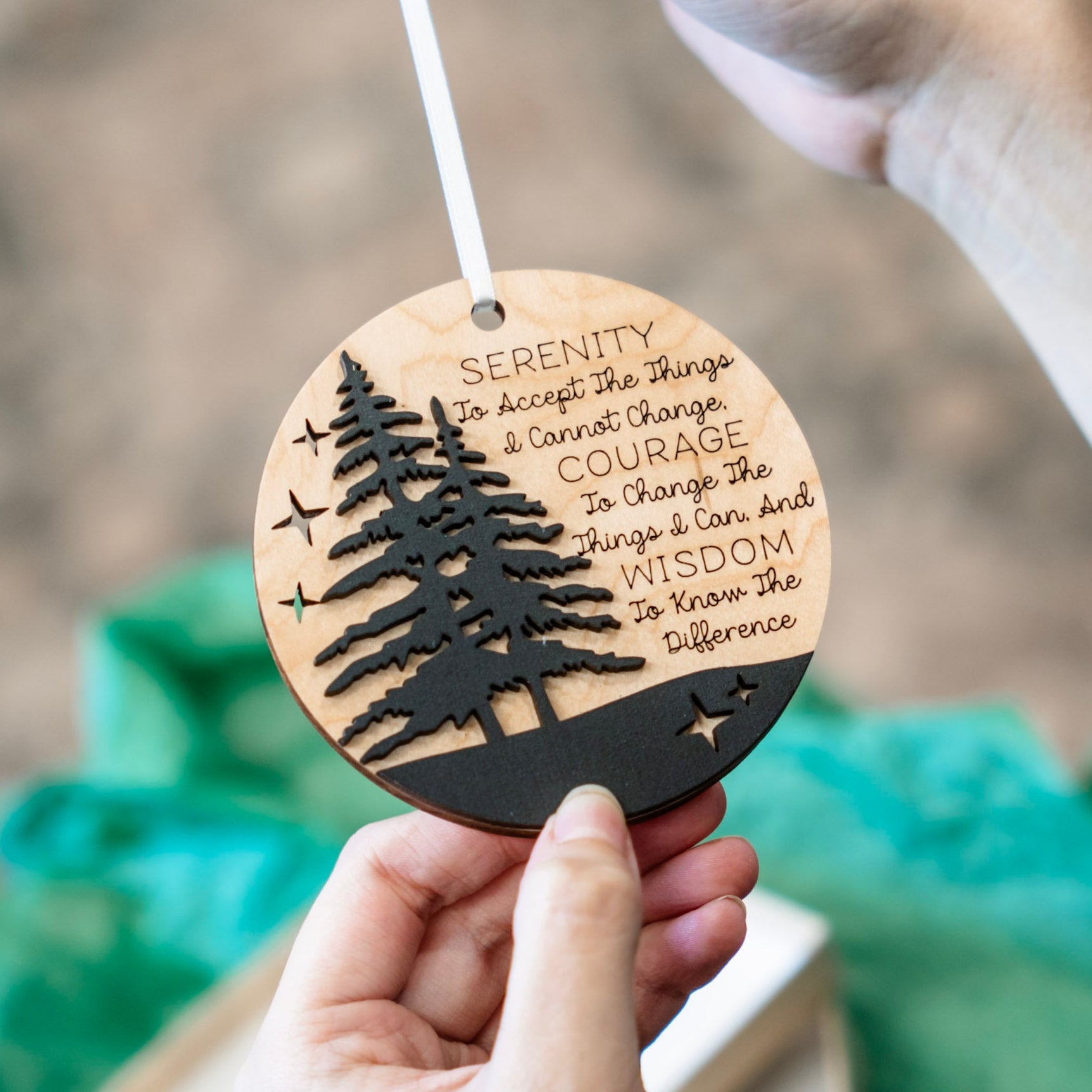 A person holds a round wooden ornament with a laser-cut tree silhouette and engraved inspirational quote. The ornament has a natural light wood finish with a black lower band and black cutout details, suspended by a white ribbon.