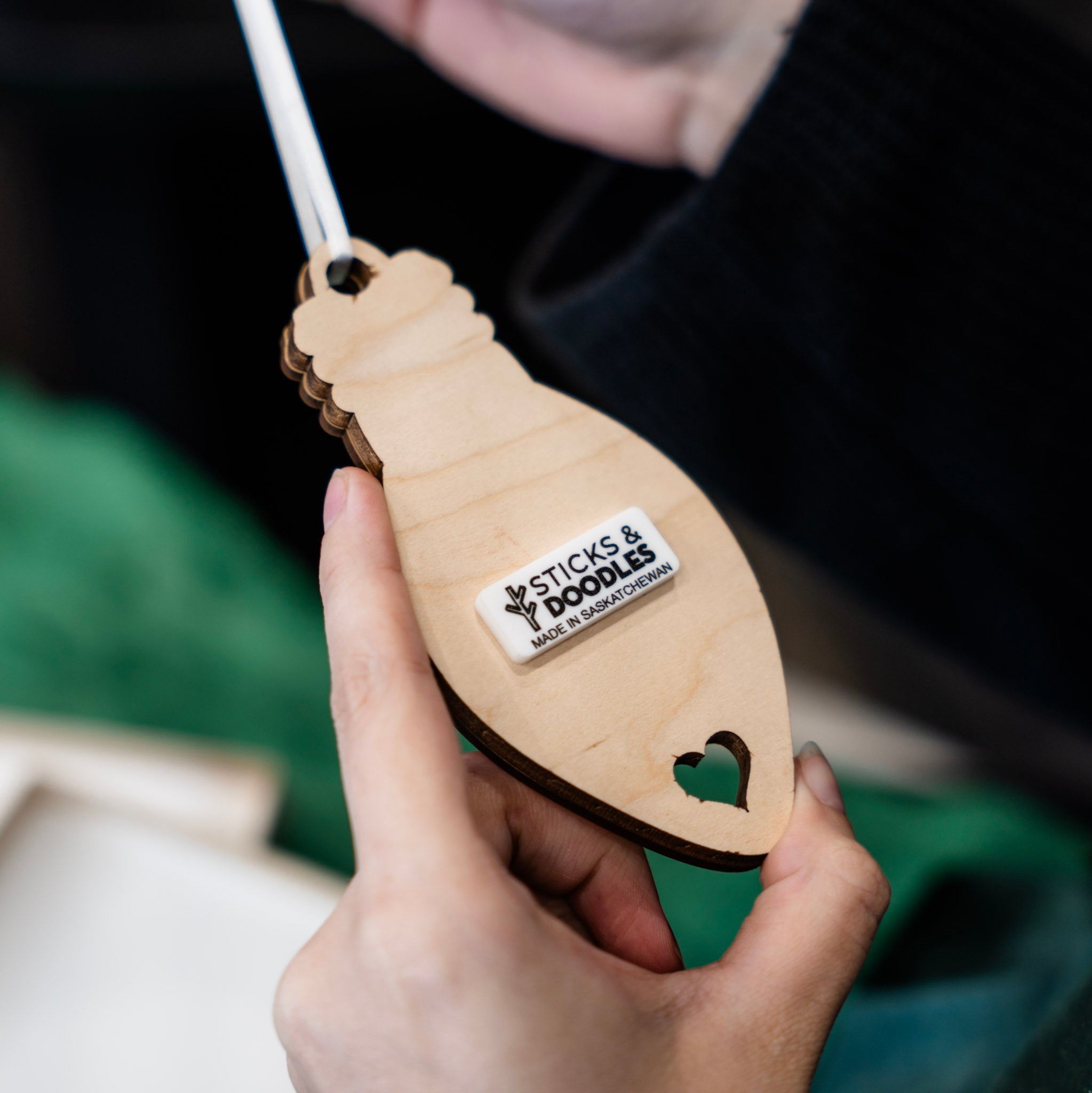 A hand holding a light-bulb-shaped wooden ornament with a heart-shaped cutout and a white sticker that reads 'STICKS & DOODLES MADE IN SASKATCHEWAN'.