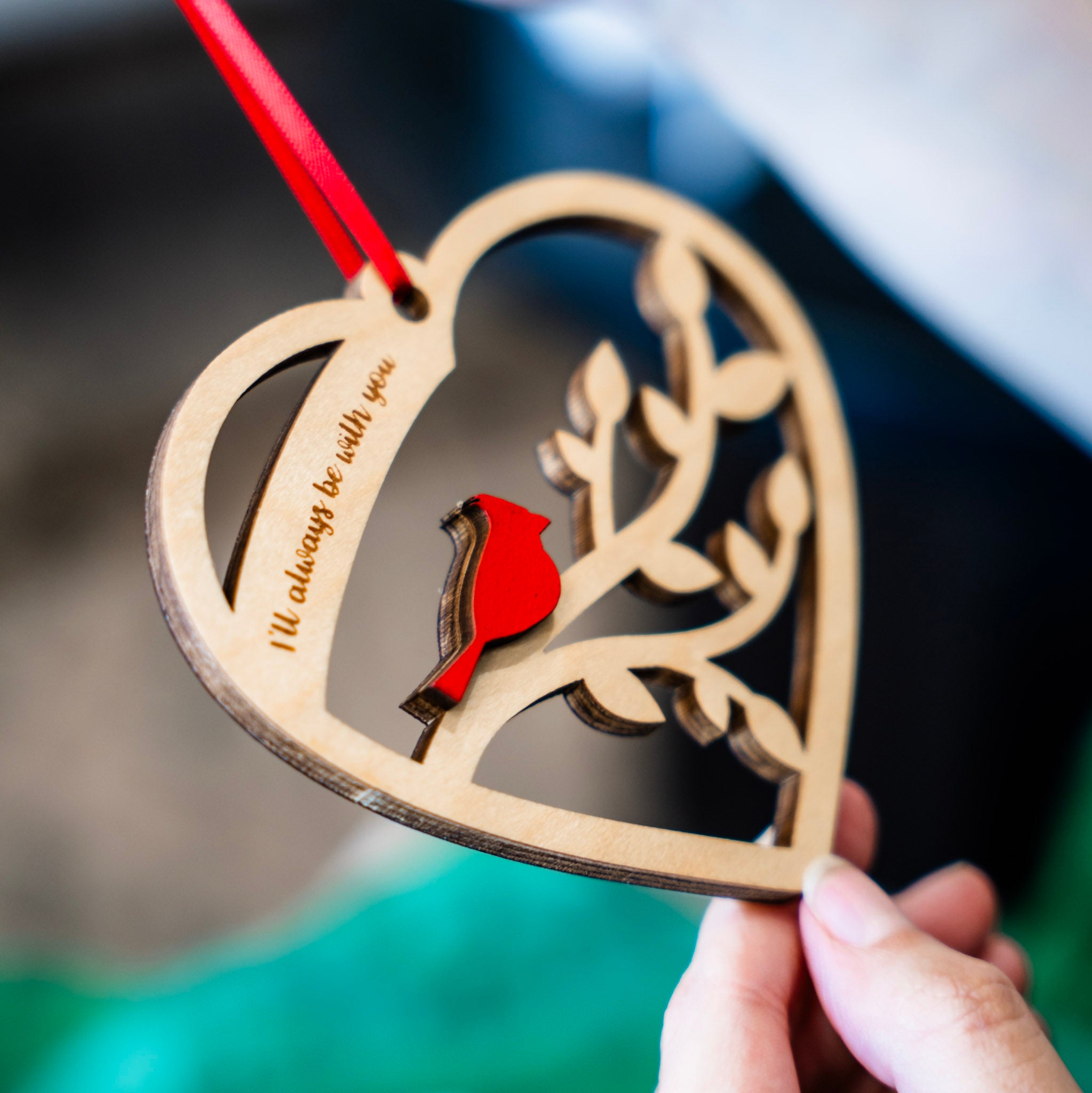 A heart-shaped wooden hanging ornament with a red ribbon, featuring a laser-cut branch design and a small red cardinal. A hand holds the ornament while another hand holds the red ribbon, with a blurred green gift bag and box in the background.