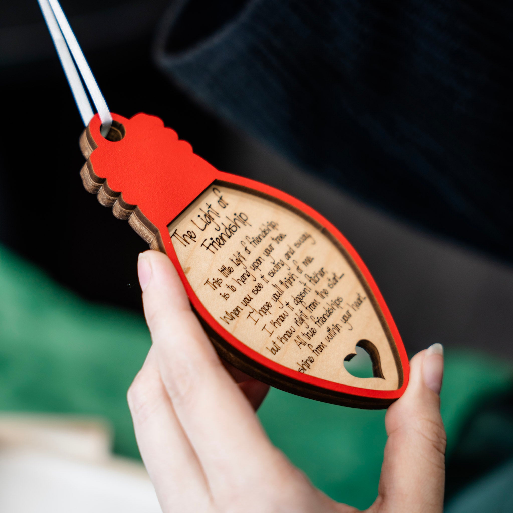 A hand holding a laser-engraved wooden lightbulb ornament with a red painted border and a white ribbon. The wooden lightbulb features a quote titled 'The Light of Friendship' and a small heart-shaped cutout.