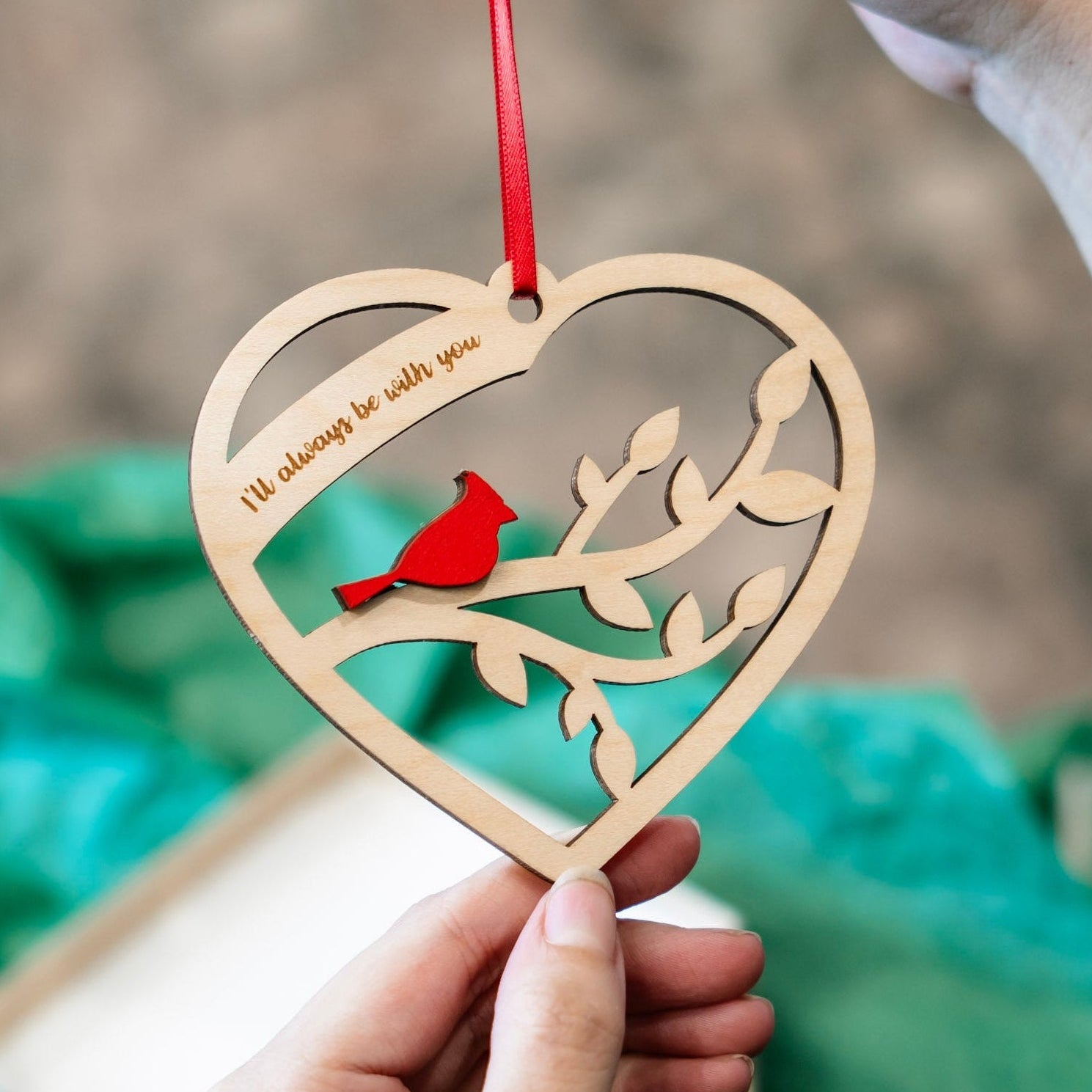 A heart-shaped wooden hanging ornament with a red ribbon, featuring a laser-cut branch design and a small red cardinal. A hand holds the ornament while another hand holds the red ribbon, with a blurred green gift bag and box in the background.