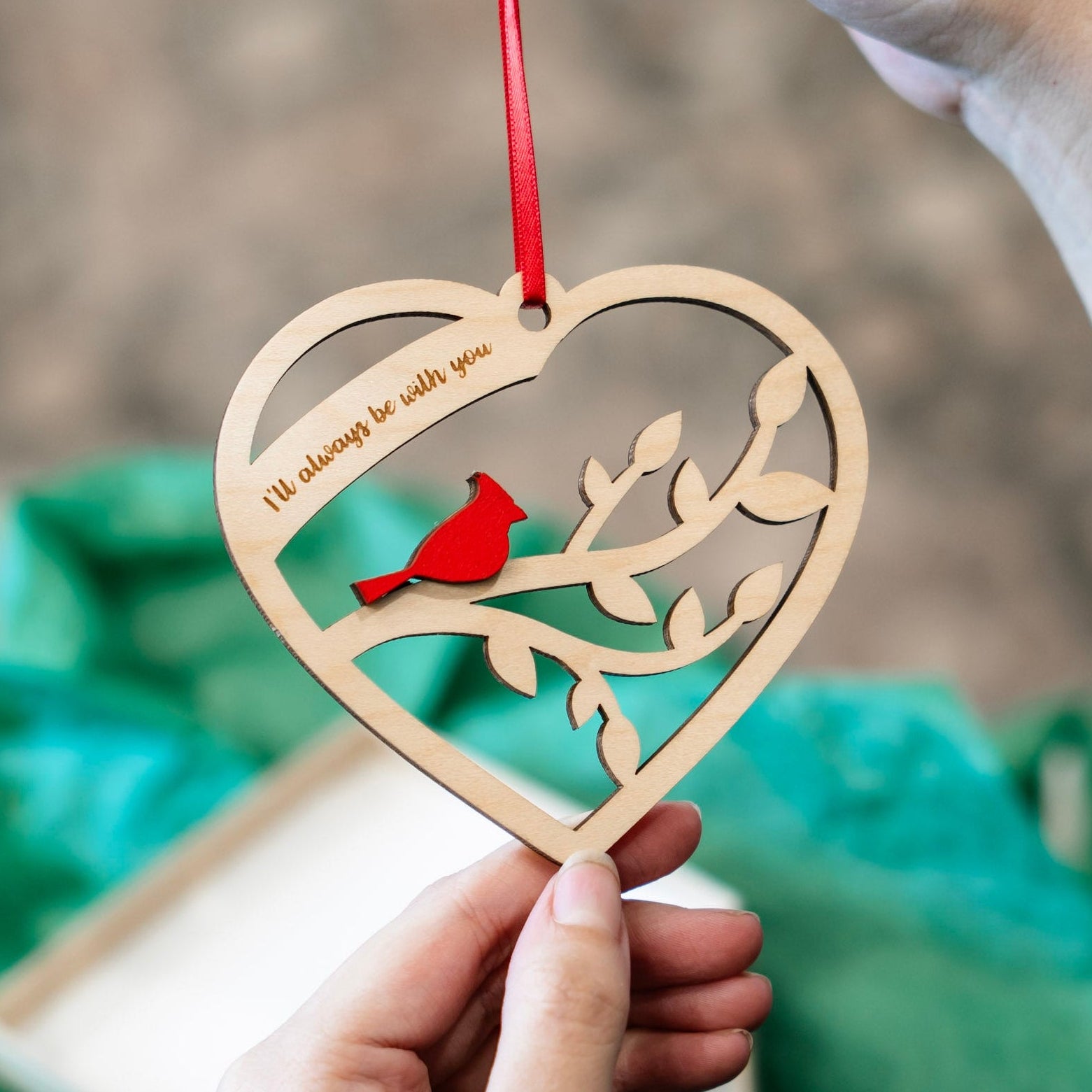 A heart-shaped wooden hanging ornament with a red ribbon, featuring a laser-cut branch design and a small red cardinal. A hand holds the ornament while another hand holds the red ribbon, with a blurred green gift bag and box in the background.