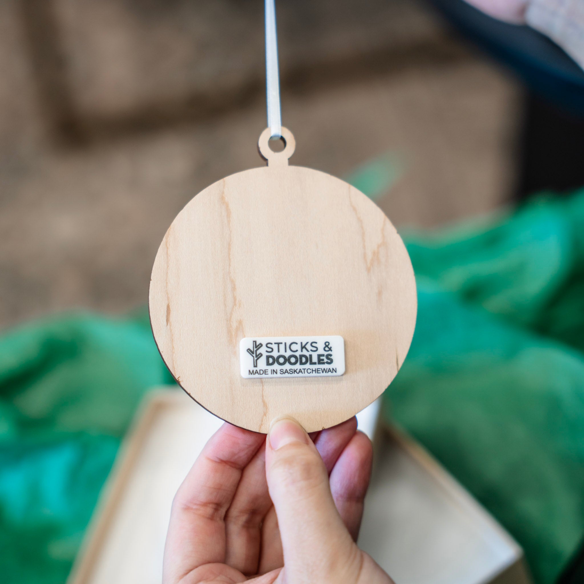A hand holding a round natural wood hanging ornament with a white string. The ornament features a small white sticker that reads 'STICKS & DOODLES MADE IN SASKATCHEWAN'.