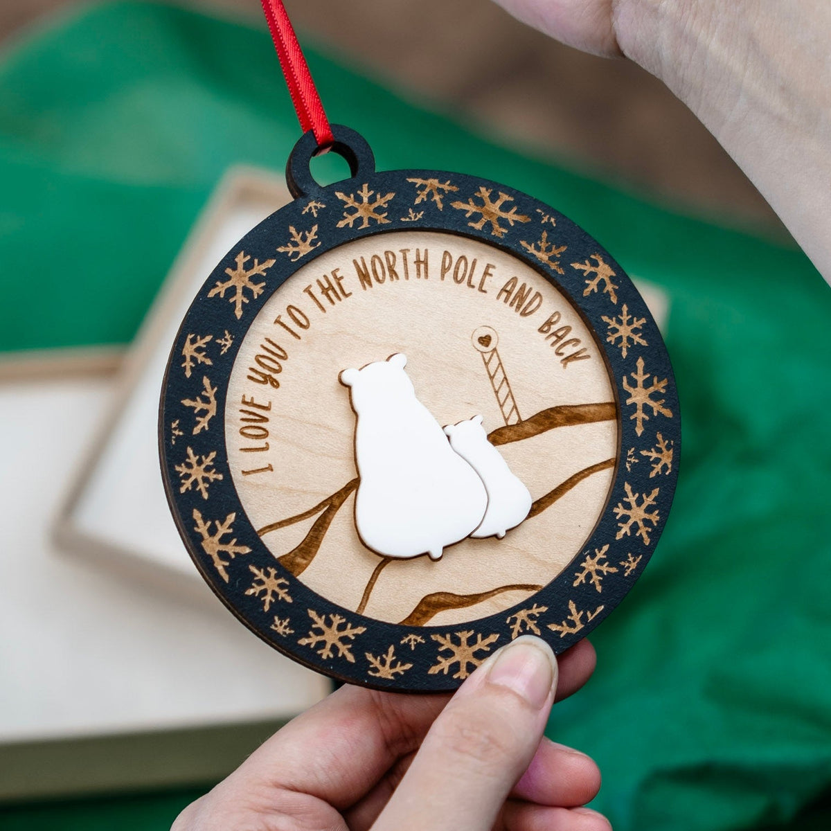 A person holds a round wooden ornament featuring two white polar bear silhouettes, engraved snow banks and pole, and the phrase 'I LOVE YOU TO THE NORTH POLE AND BACK' on a red hanging ribbon. A green tissue paper backdrop and part of a gift box are visible in the background.