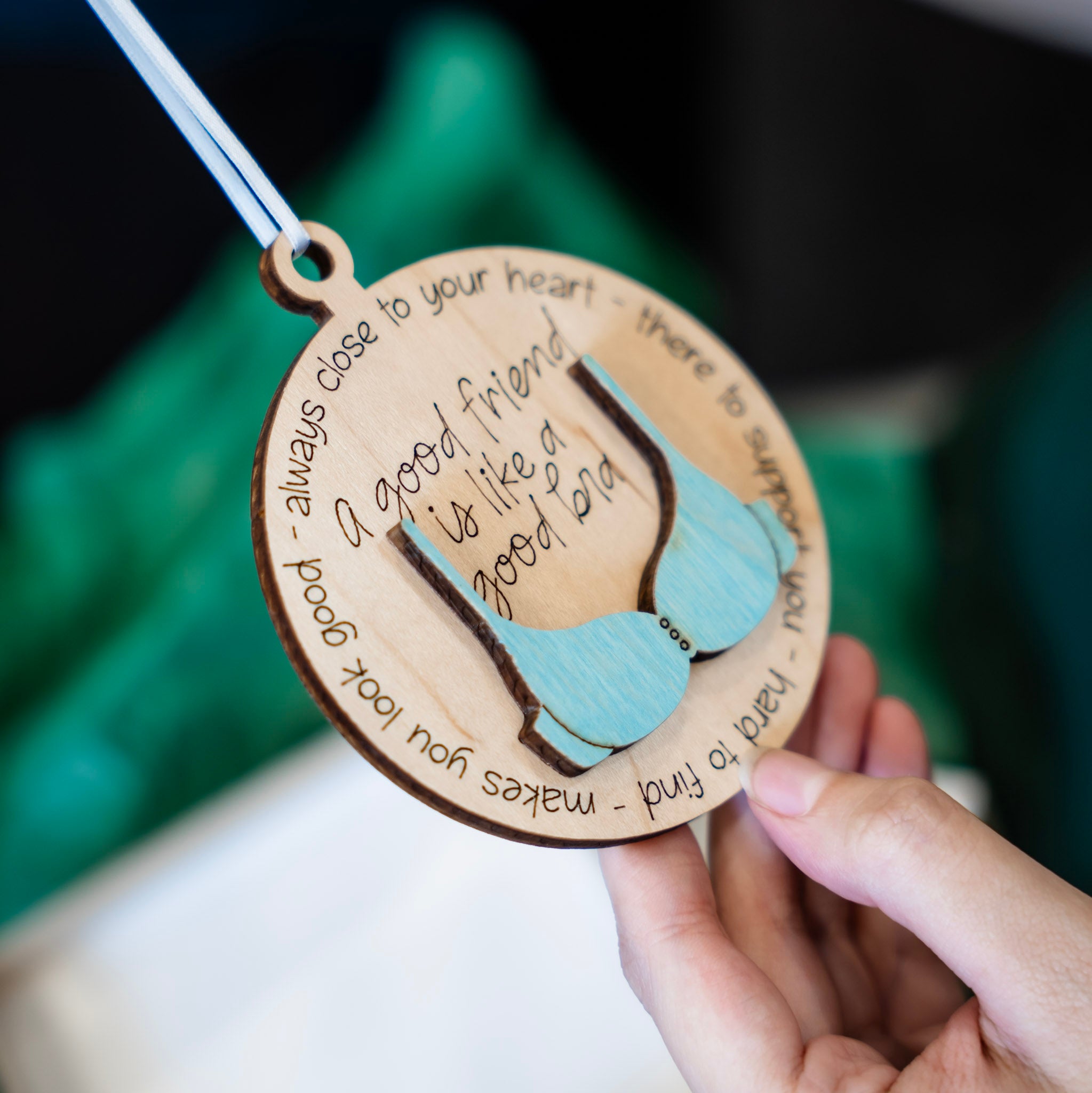 A hand holds a round wooden ornament engraved with a 3D bra and the quote 'a good friend is like a good bra' surrounded by supportive words. The background is softly blurred with green tissue paper and a neutral indoor setting.