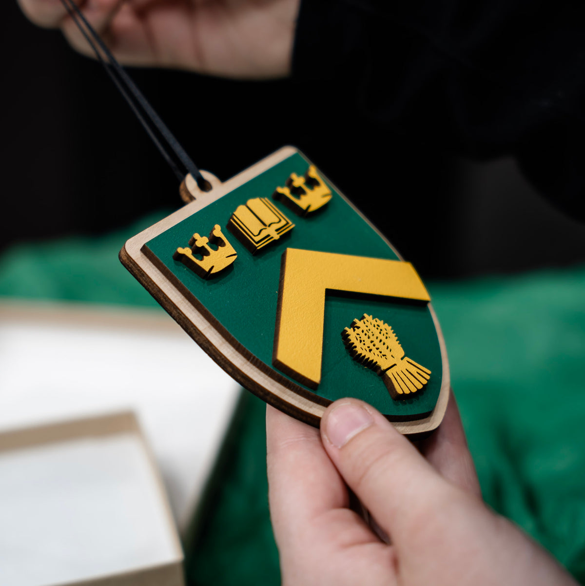 A shield-shaped wooden logo ornament of the University of Regina, featuring a green and yellow design with two gold crowns, an open book, and a bundle of wheat, held by a hand against a green fabric background with a gift box. 