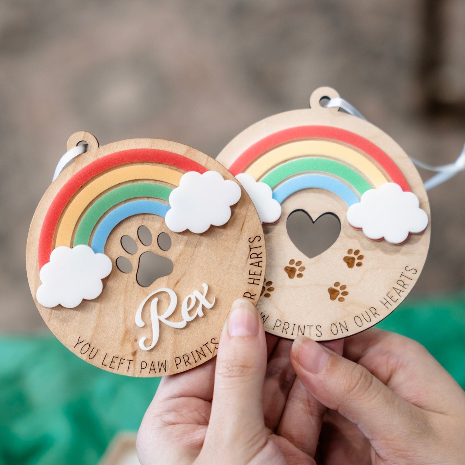 Two personalized wooden pet memorial ornaments held in hands, featuring colorful rainbow designs, white clouds, paw print cutouts, and engraved text including 'Rex' and 'YOU LEFT PAW PRINTS ON OUR HEARTS'.