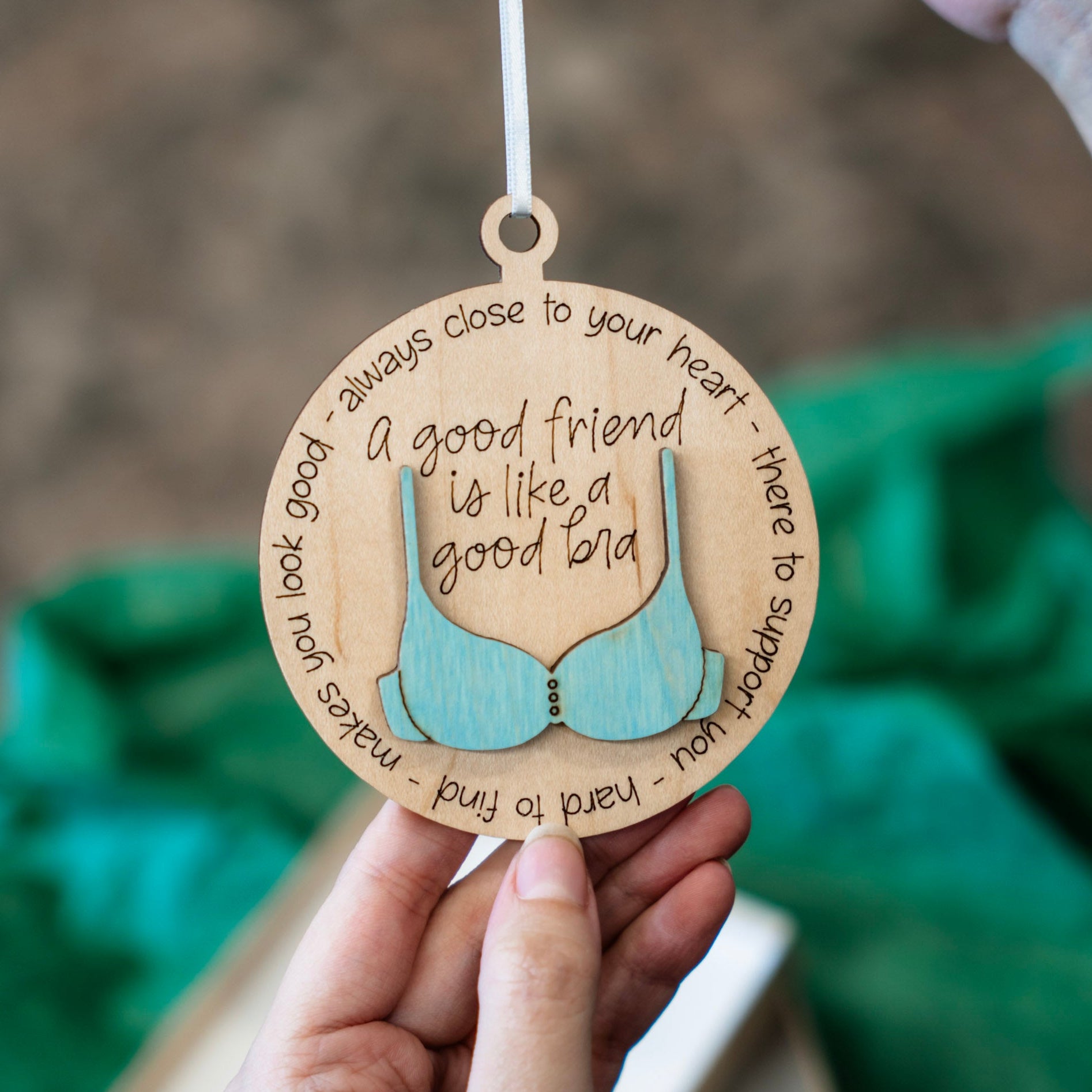 A hand holds a round wooden ornament engraved with a 3D bra and the quote 'a good friend is like a good bra' surrounded by supportive words. The background is softly blurred with green tissue paper and a neutral indoor setting.