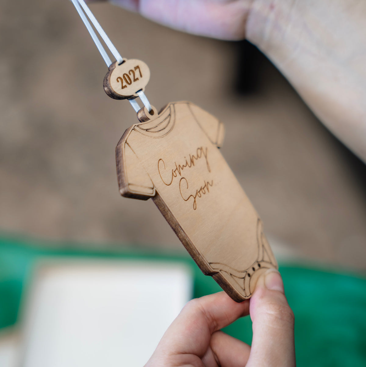 A personalized wooden baby bodysuit ornament held by a hand, featuring the engraved text 'Cetaincy Gooch' and a hanging tag that reads '2027'.