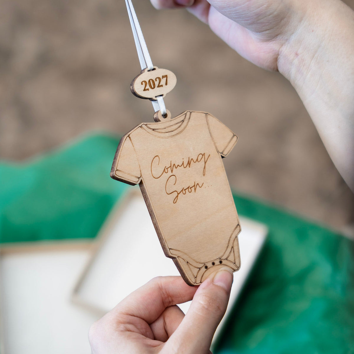 A close-up of hands holding a wooden baby bodysuit ornament engraved with 'Coming Soon' and '2027', with a green tissue and gift box blurred in the background.