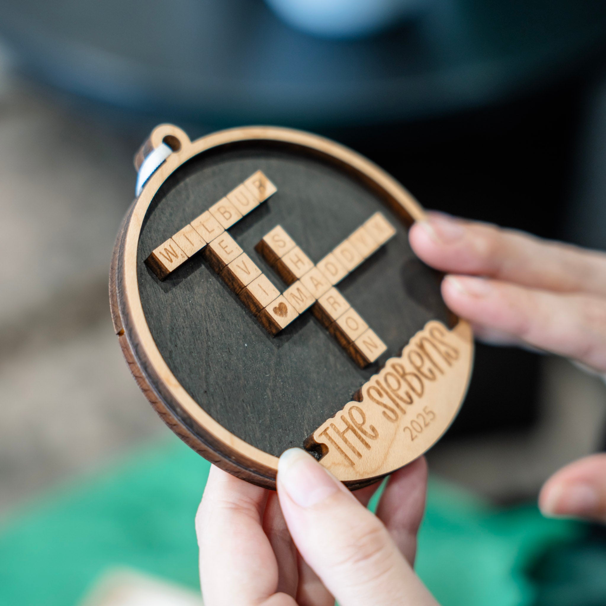 A close-up of a round wooden keepsake ornament being held by hands. The ornament features a dark wood center with a wooden letter grid puzzle spelling 'THE SIEBENS 2025' and a light wood border.