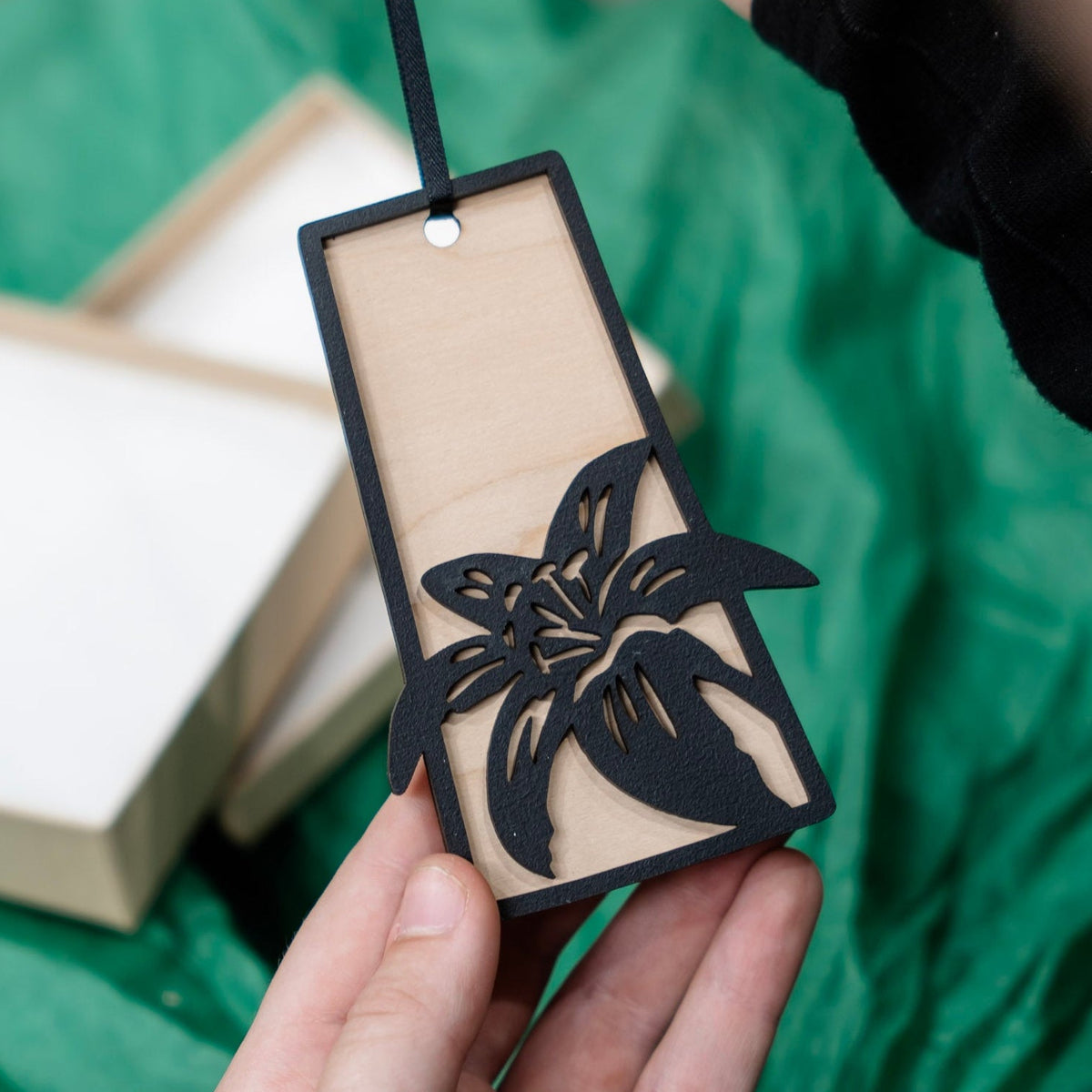 A hand holds a Saskatchewan outline wooden ornament with a black laser-cut flower lily design, set against a green fabric background with gift boxes and tissue paper blurred in the background.