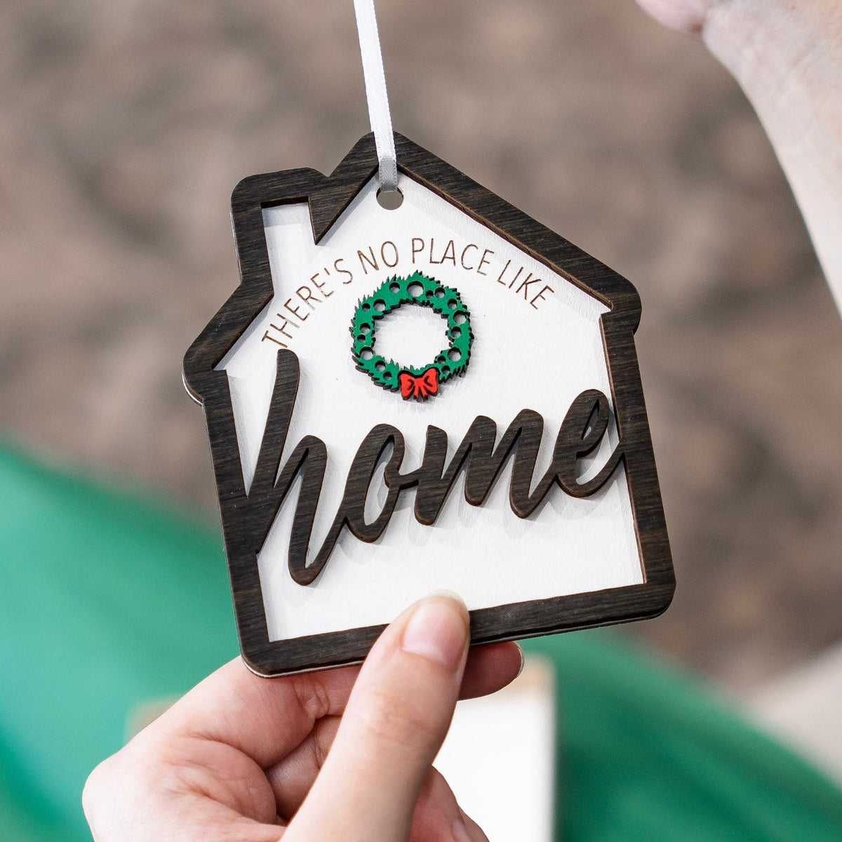 A close-up of a hand holding a house-shaped wooden ornament with the word 'home' in script and a small green wreath with a red bow at the top center. 