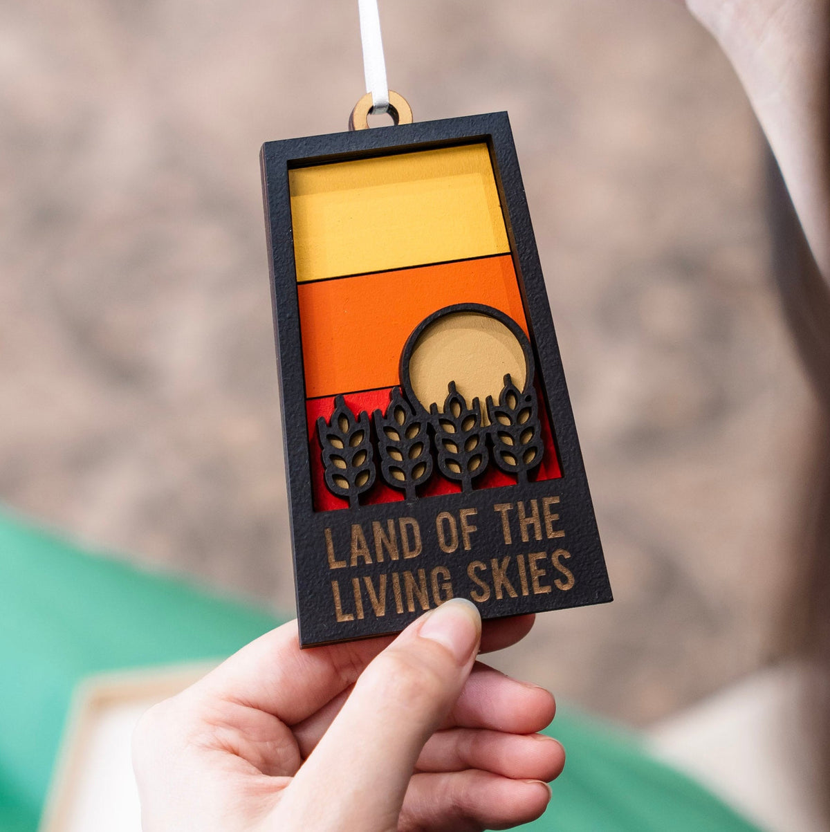 A hand holds a rectangular black wooden ornament with a white hanging string. The ornament features a sunset-style design with yellow, orange, and red geometric blocks, a circular gold-toned element, black wheat motifs, and the text 'LAND OF THE LIVING SKIES'.