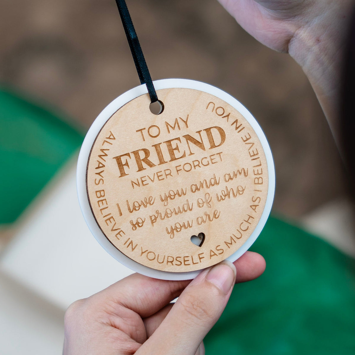 A close-up of a person holding a round wooden engraved ornament. The ornament features engraved text reading 'TO MY FRIEND NEVER FORGET I love you and am so proud of who you are. ALWAYS BELIEVE IN YOURSELF AS MUCH AS I BELIEVE IN YOU'.