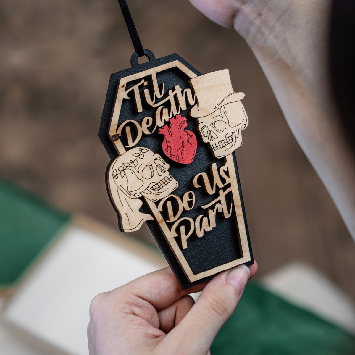 A person holds a coffin-shaped wooden ornament with the phrase 'Til Death Do Us Part' in laser-cut wood, featuring a red anatomical heart and skull bride and groom details, against a blurred background.