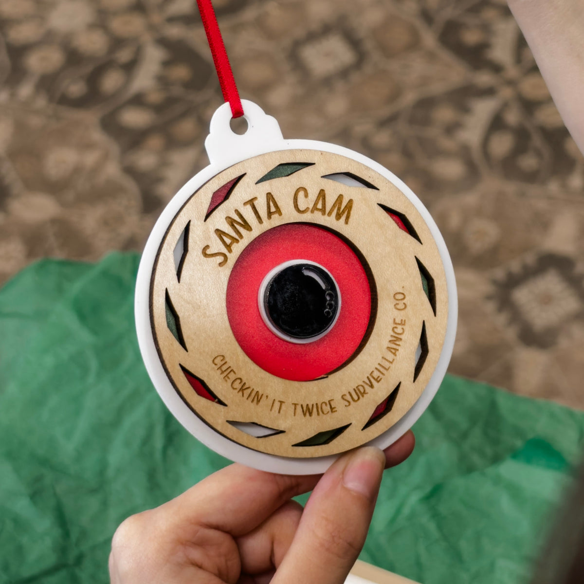 A hand holding a round wooden Santa Cam ornament with a red hanging ribbon. The ornament features a natural wood face with a red and black camera lens design in the center, surrounded by the words 'SANTA CAM' and 'CHECKIN' IT TWICE SURVEILLANCE CO.