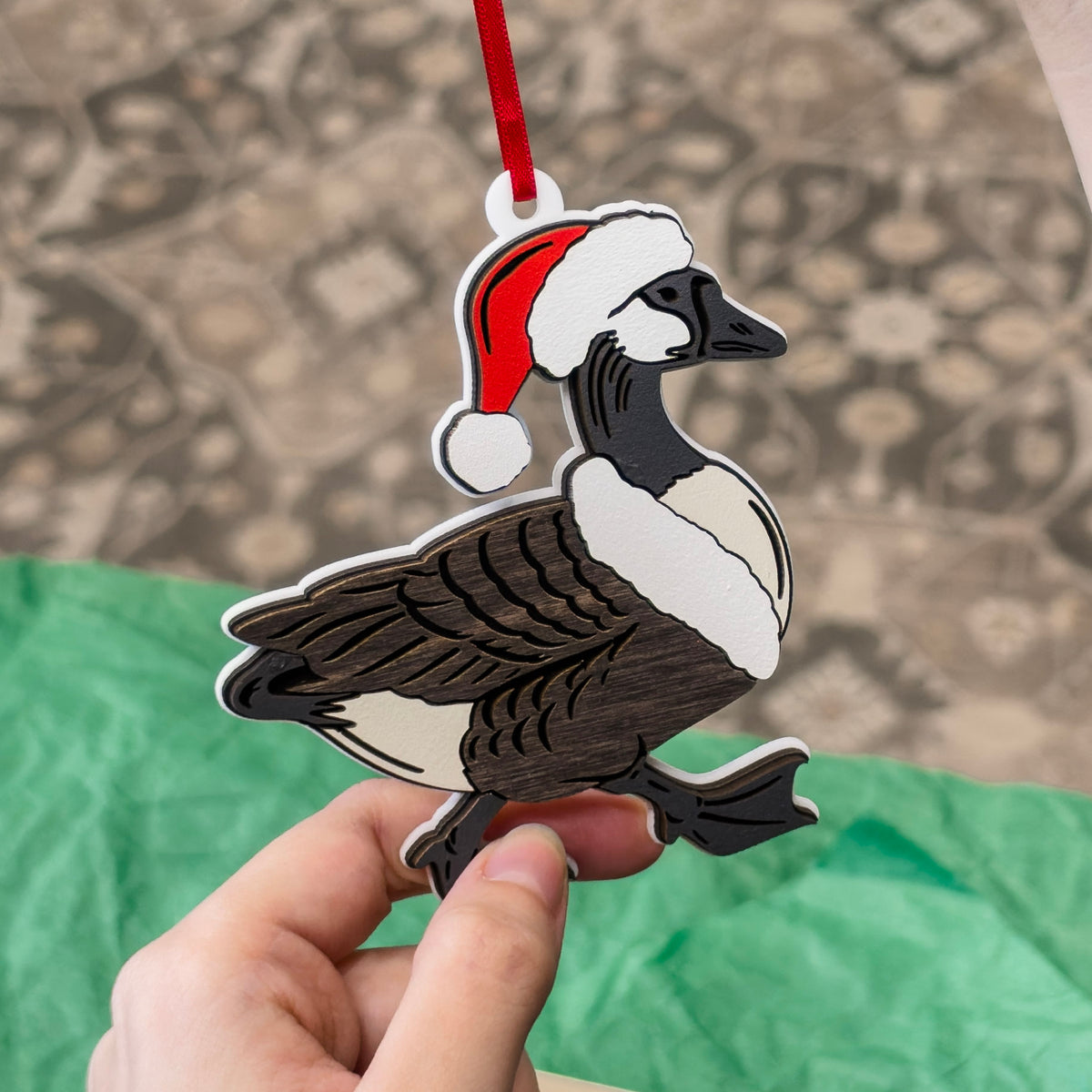 A hand holding a Christmas ornament shaped like a Canada goose wearing a red Santa hat, with a red hanging ribbon, against a blurred carpet background and green tissue paper.