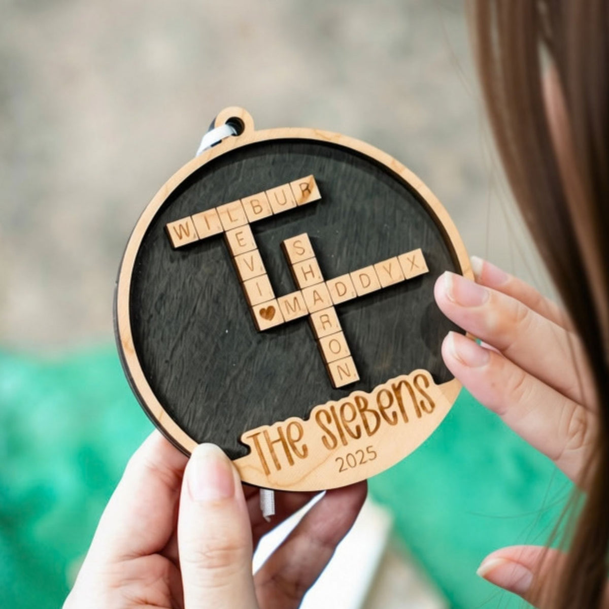 A personalized round wooden ornament held by two hands, featuring a dark wood center with a crossword-style arrangement of Scrabble tiles with four different names, plus the engraved text 'The Siebens' and '2025' on a light wood border.