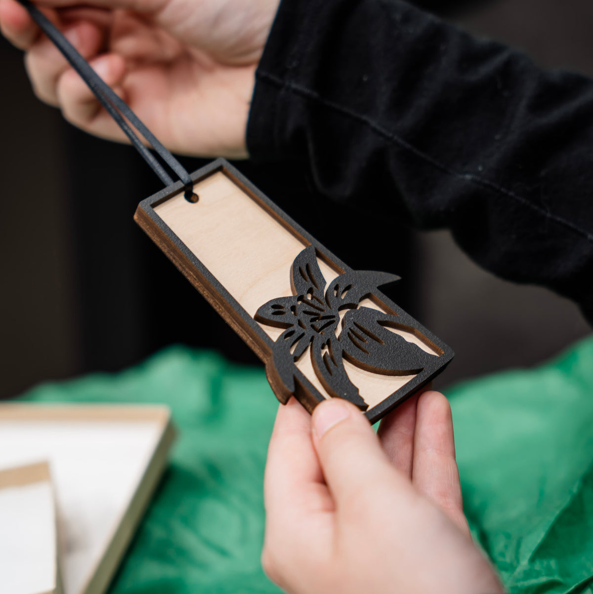 A close up of hands holding a Saskatchewan outline wooden ornament with a black laser-cut flower lily design, set against a green fabric background with gift boxes and tissue paper blurred in the background.