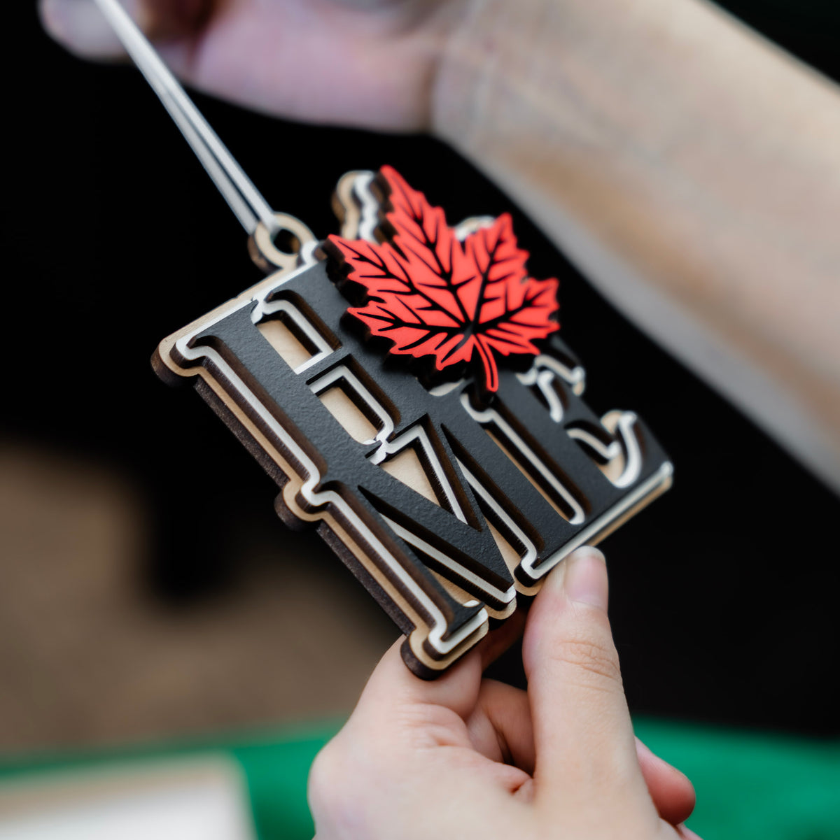 A close-up of a hand holding a wooden 'HOME' ornament with a red maple leaf, with another hand holding the white ribbon at the top, set against a green fabric background.