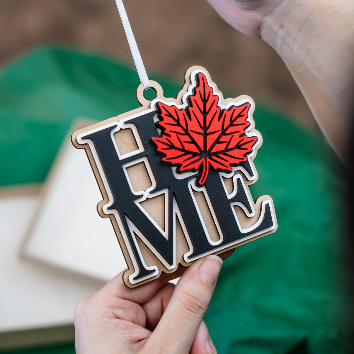 A close-up of a hand holding a wooden 'HOME' ornament with a red maple leaf, with another hand holding the white ribbon at the top, set against a green fabric background.