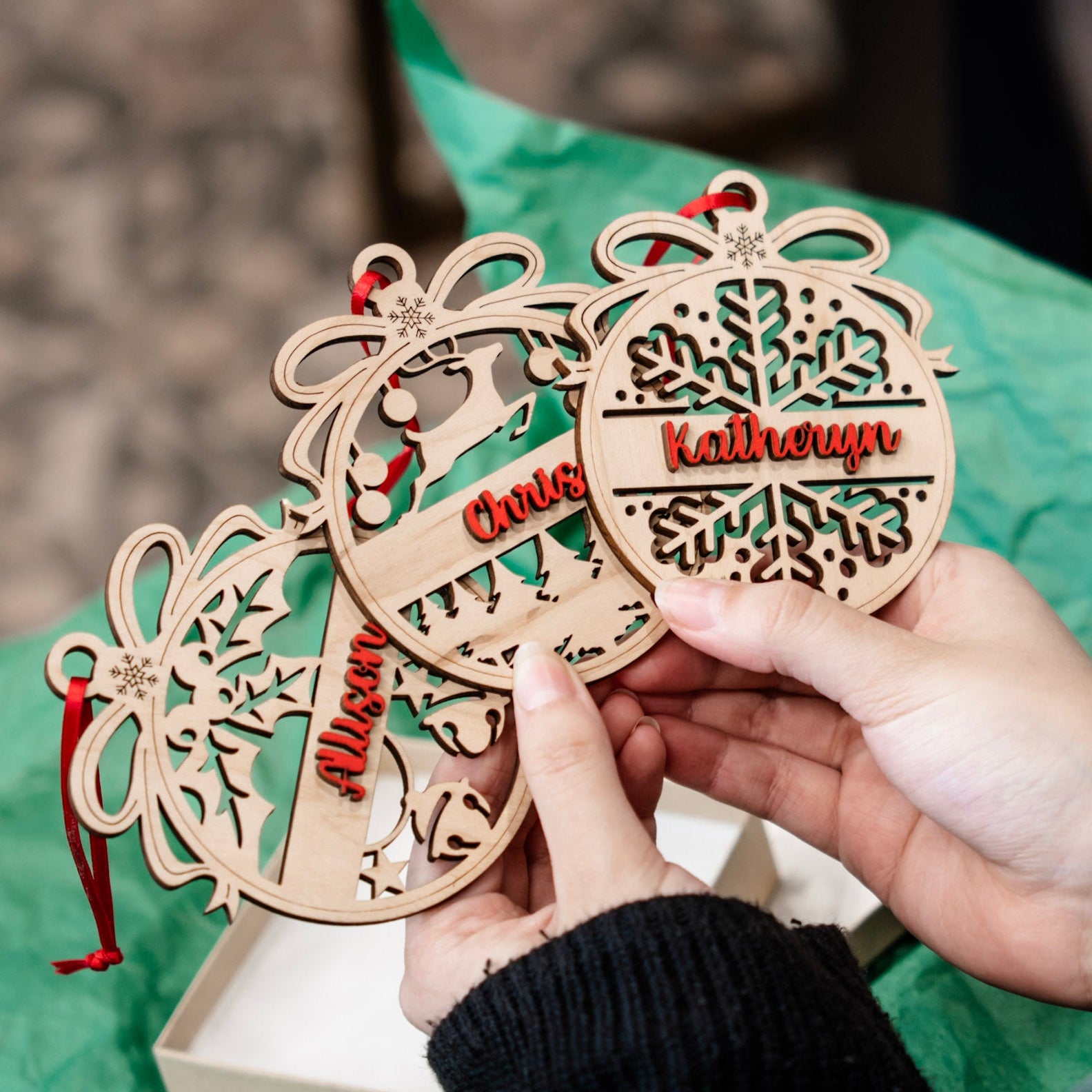 Three personalized wooden Christmas ornaments held by hands over a green tissue paper and gift box. The ornaments are laser-cut with holiday designs and names including 'Kathleen' and 'Chris'.