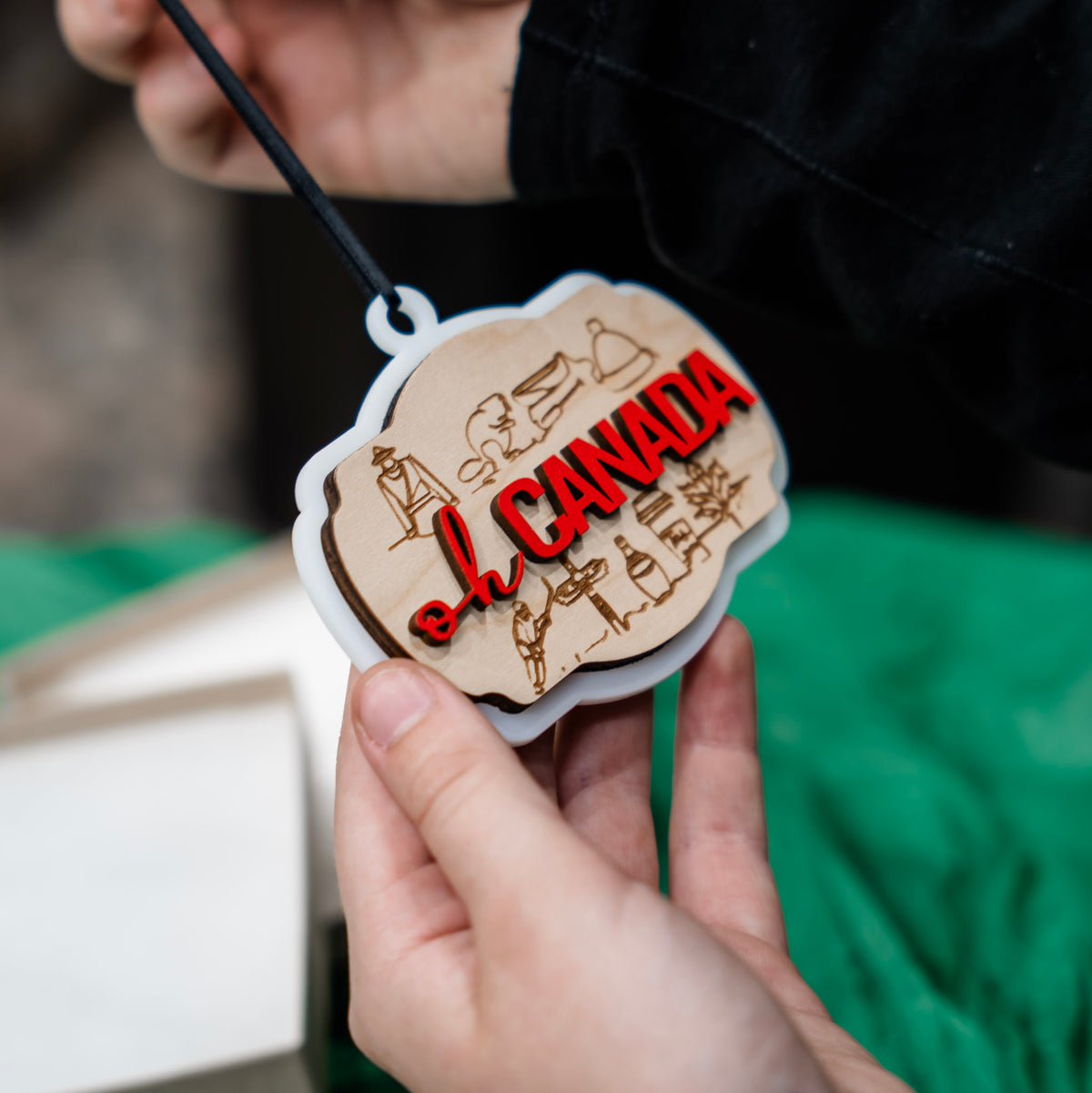 A hand holds a small wooden ornament with acrylic borders that says oh Canada with many icons of Candian things. The ornament hangs from a black string, with a green tissue paper backdrop and a gift box.