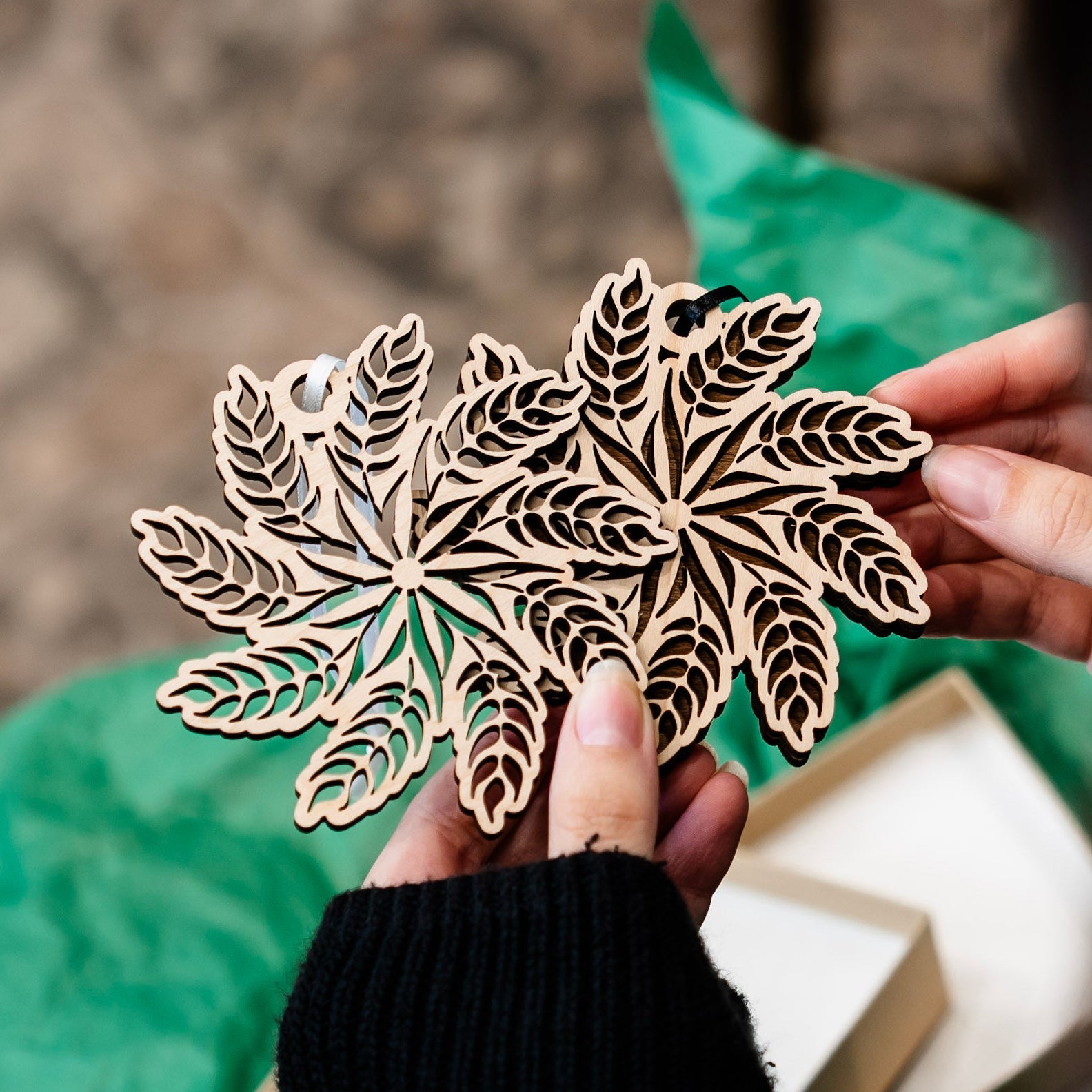 A person holds two laser-cut wooden ornament shaped like a snowflake made from wheat, one with a single light wood layer and the other with a darker background layer behind the light wood. The ornament is shown close up against a soft-focus background with green tissue paper and a light box.