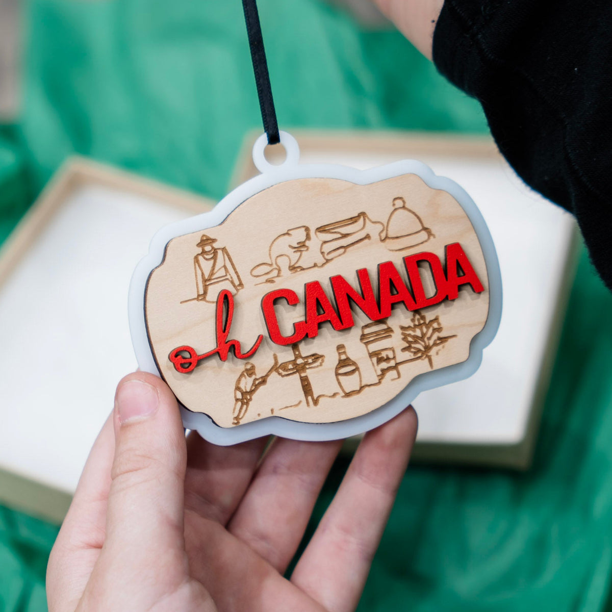 A hand holds a small wooden ornament that says oh canada with many icons of Candian things. The ornament hangs from a black string, with a green tissue paper backdrop and a gift box.