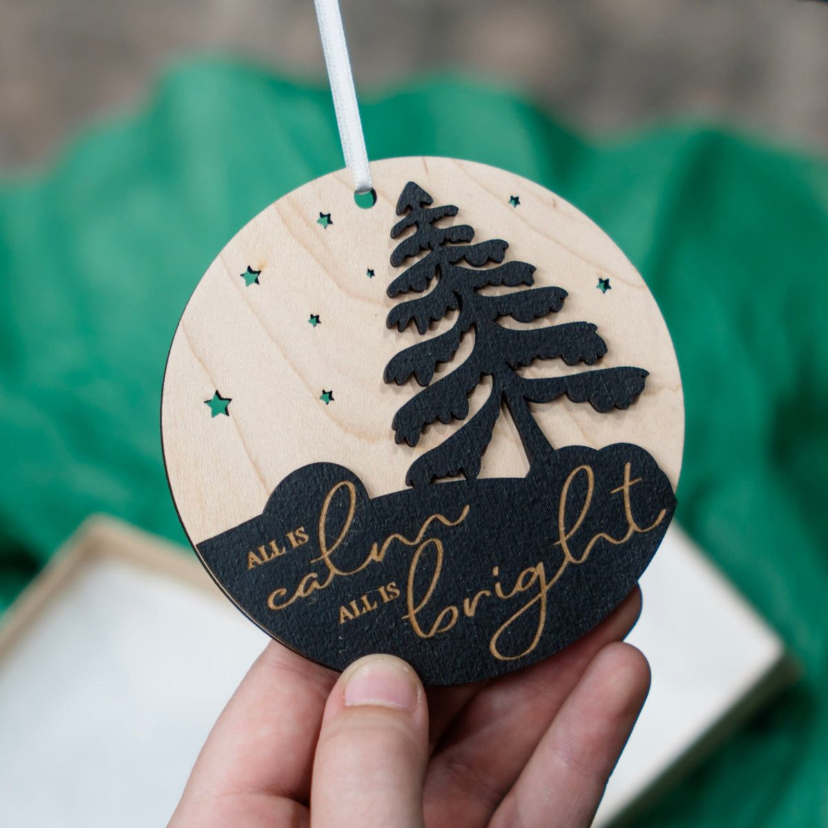A hand holds a round wooden Christmas ornament featuring a laser-cut tree, stars, and the words 'All is calm all is bright' in gold on a black section, with a green fabric background.