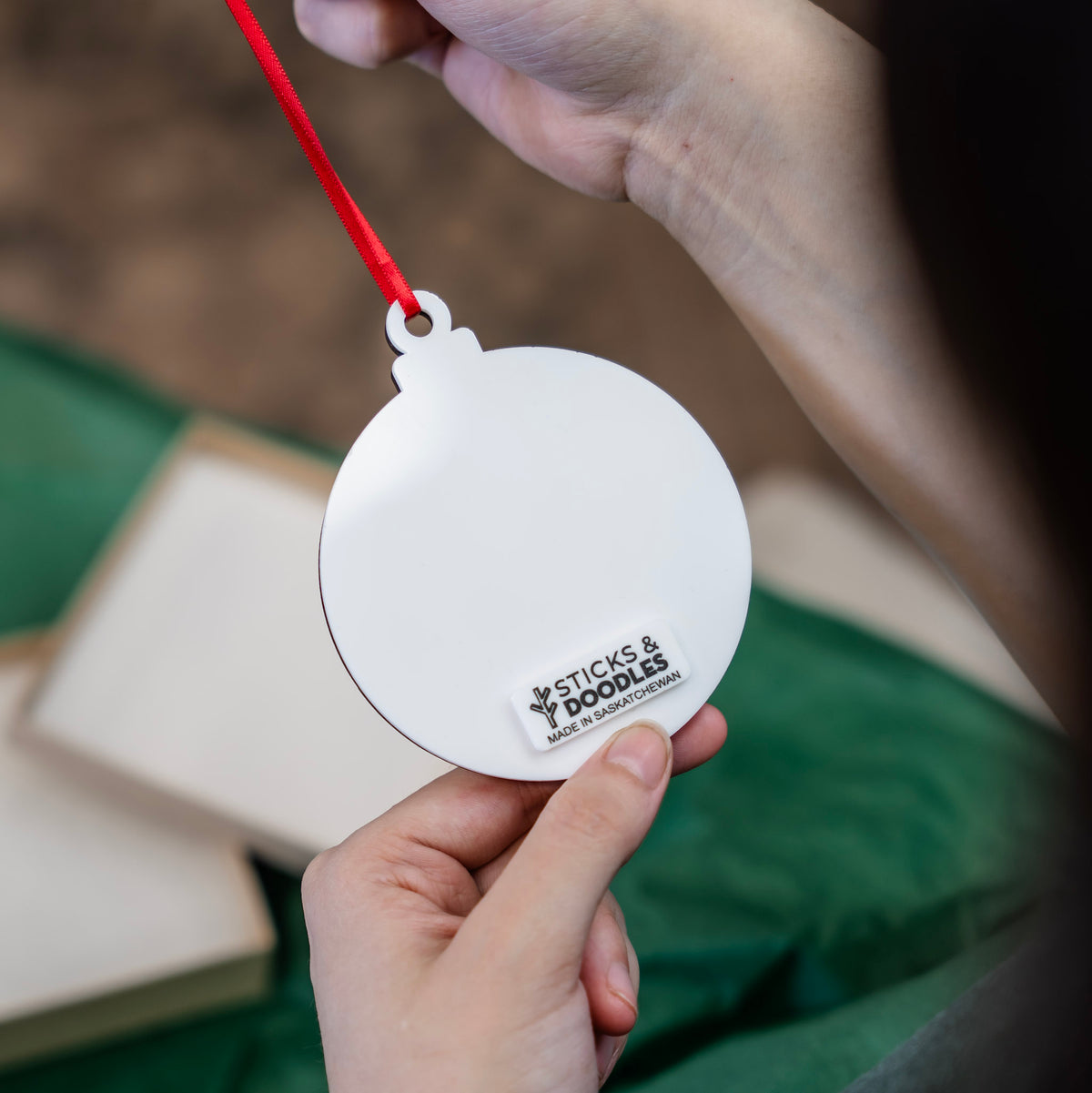 Hands hold the back of the ornament with a white acrylic backer and a tag that reads Sticks & Doodles Made in Saskatchwewan. The ornament hangs with a red ribbon.