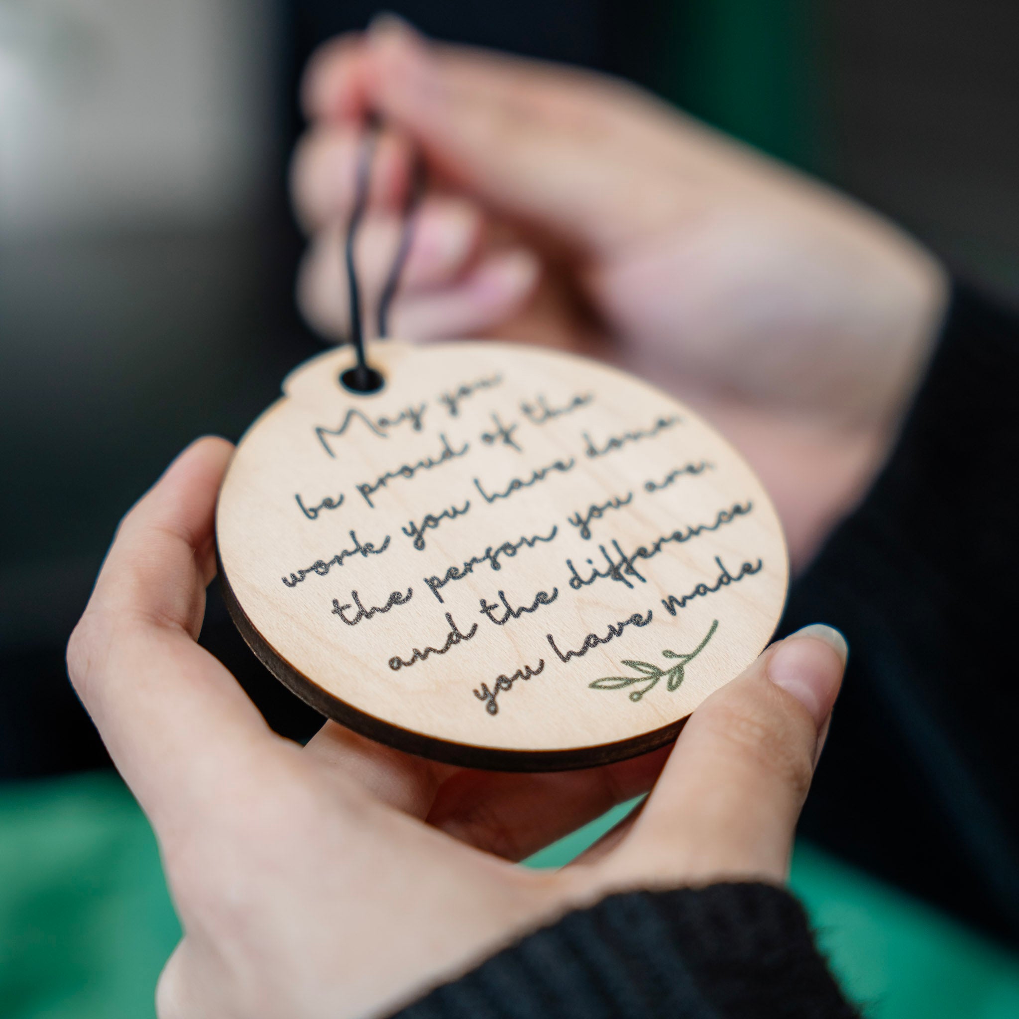 A person holding a round wooden keepsake tag engraved with a motivational quote in black script and a small green leaf motif. The tag is suspended from a black cord, with the person's hands holding it against a blurred dark background.