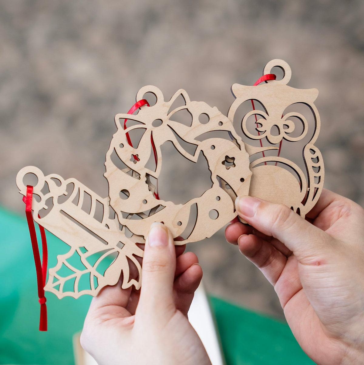 A person's hands holding a laser-cut wooden Christmas ornament set featuring a wreath, candy cane, and owl, with red hanging ribbons, against a blurred background.
