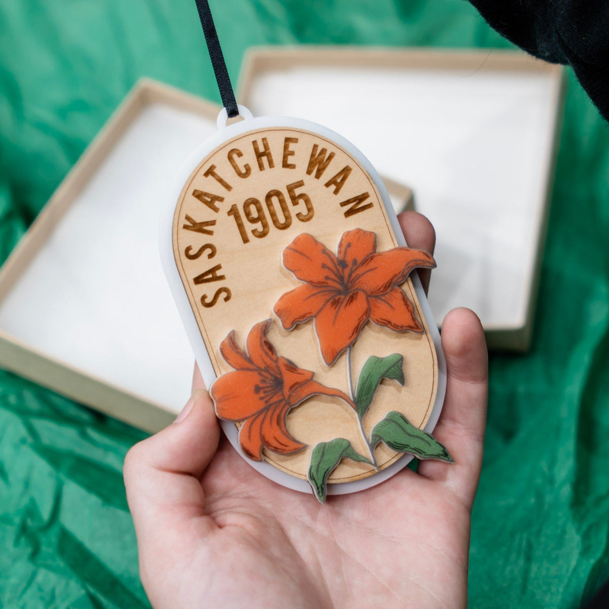 A hand holds a three layer wooden and acrylic ornament engraved with 'SASKATCHEWAN 1905' and two orange lilies on top . An open gift box and green wrapping paper are visible in the background.