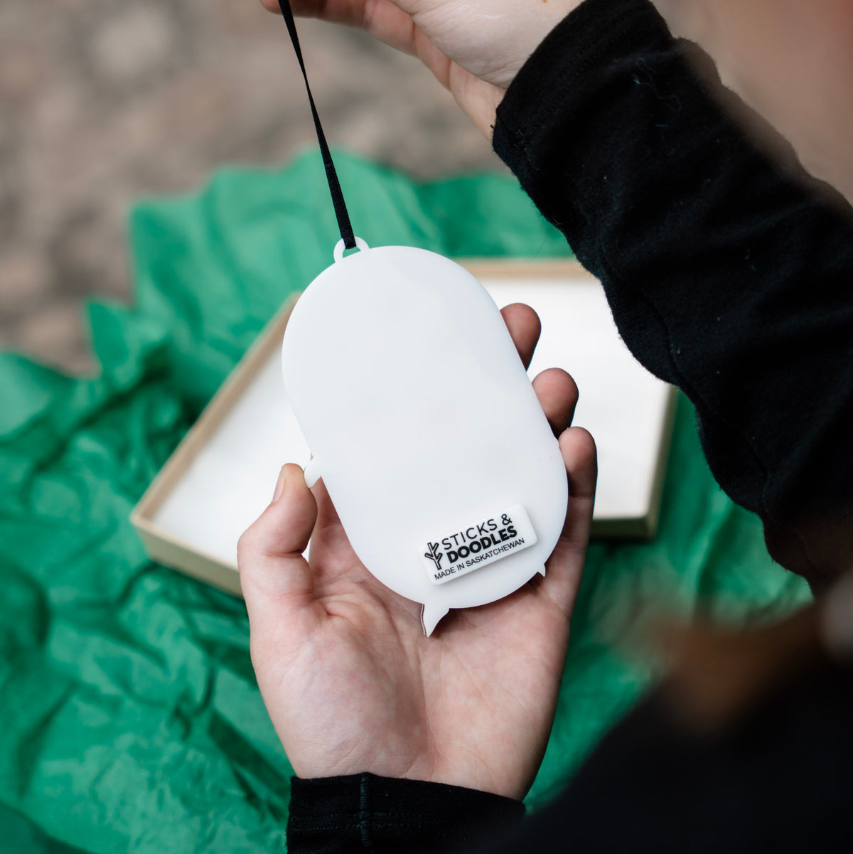 The back view of the oval shaped ornament that is made of acrylic, with a tag that reads STICKS & DOODLES MADE IN SASKATCHEWAN.