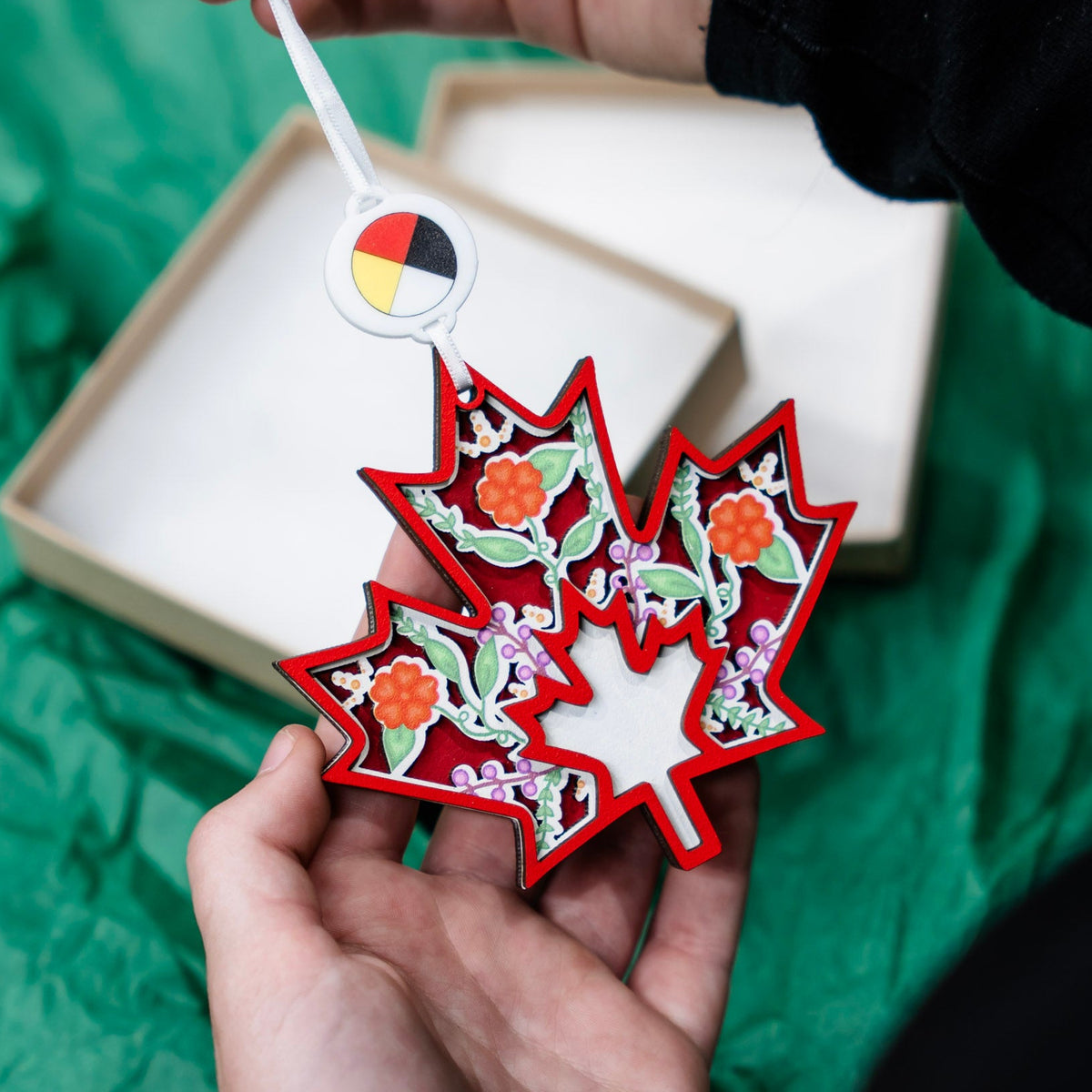 A hand holds a red maple leaf ornament decorated with layered floral and leaf patterns, while the white ribbon holds a small circular tag above it. An open beige gift box and crumpled green tissue paper are visible in the background.