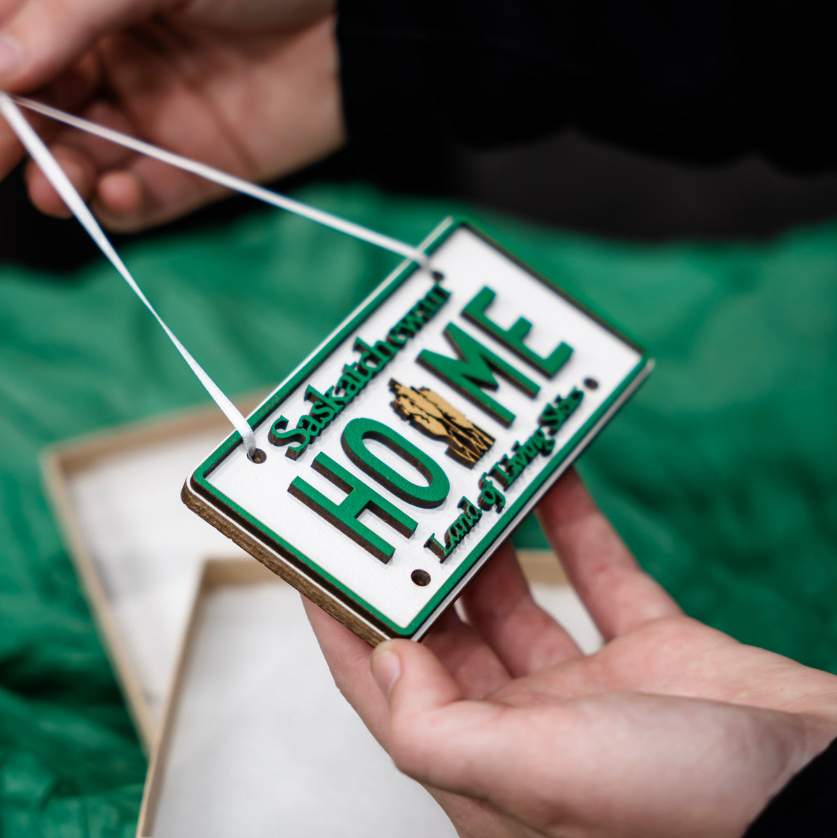 A close-up of hands holding a rectangular ornament shaped like a white license plate with green border and the words 'Shaktinoven Home Land of the Free.' A white string is being held up by another hand.