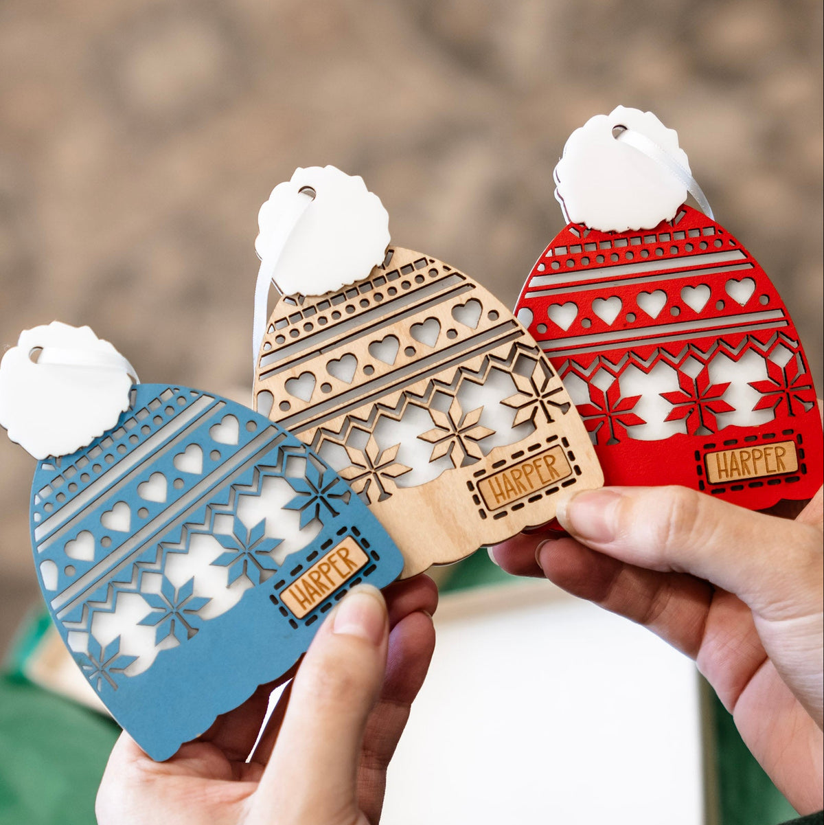 Three laser-cut wooden Christmas ornament shaped like knit winter Toques are held in two hands. The ornaments are in blue, natural wood, and red, each with a white faux-fur pom-pom and a small wooden tag that spells out 'Harper'.