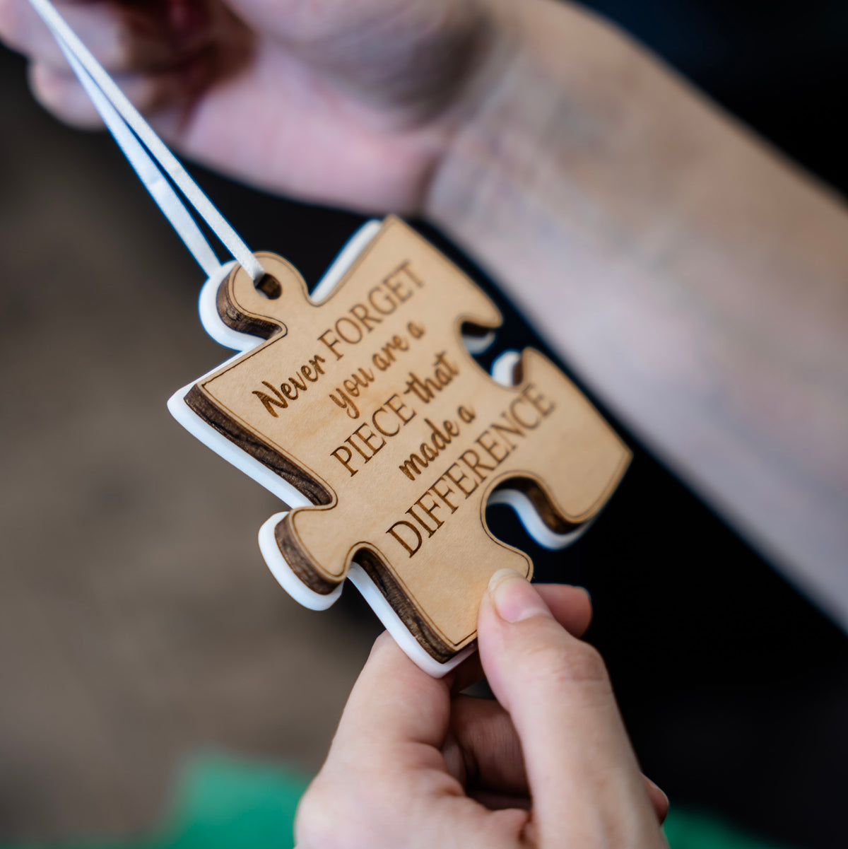 A person holds a wooden puzzle-shaped ornament engraved with the quote 'Never FORGET you are a PIECE that made a DIFFERENCE' on a white ribbon, with white acrylic backer with a blurred green gift bag in the background.