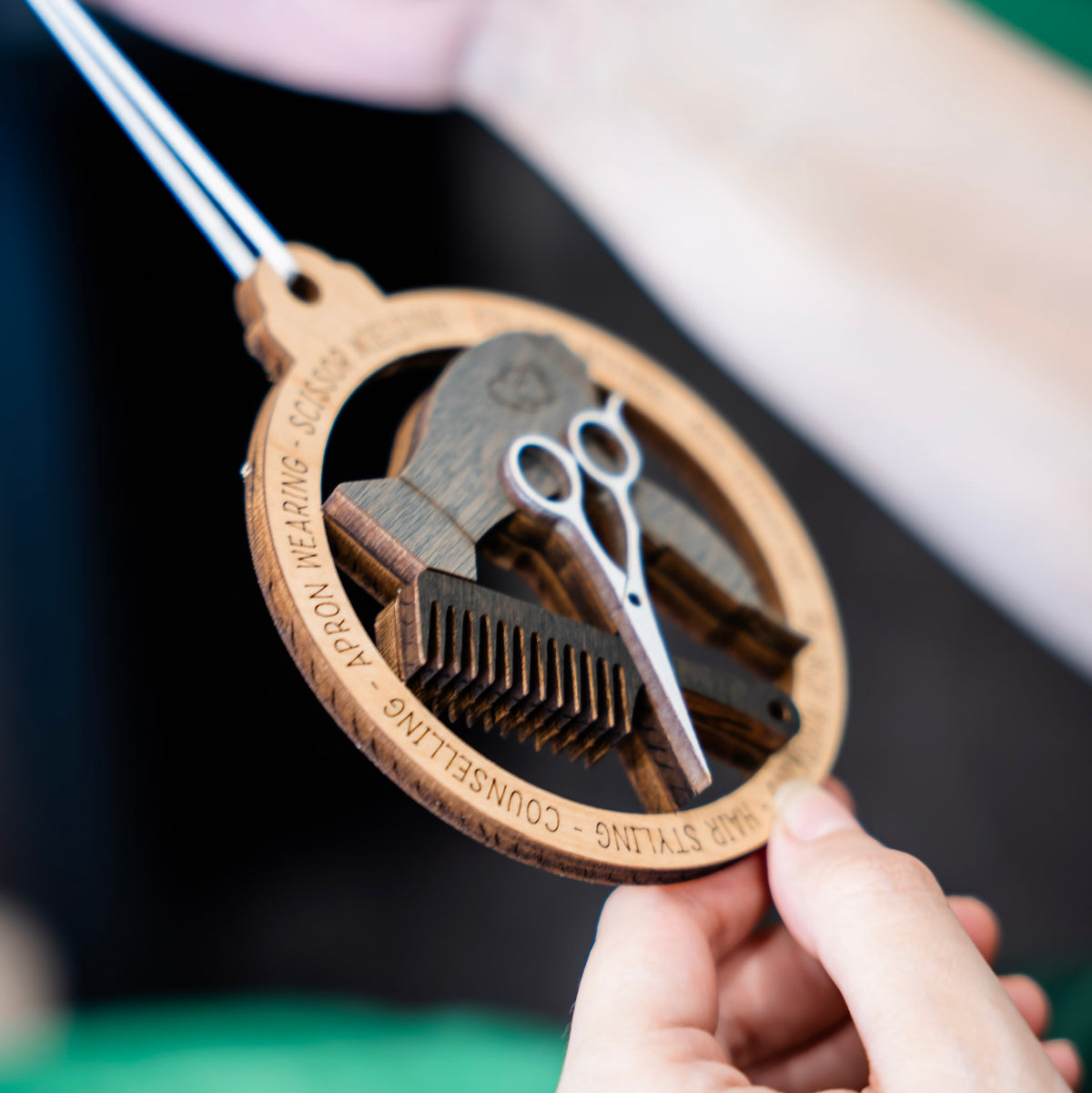 A close-up of a hand holding a circular wooden hair stylist ornament with scissors, comb, and curling iron cutouts. The wood is engraved with hair styling terms, and hangs with a white ribbon.