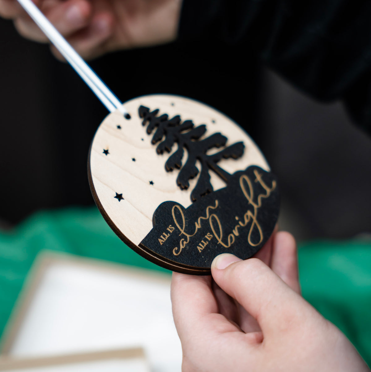 A close-up of a hand holding a round wooden ornament with a black tree silhouette, engraved text reading 'all is calm all is bright,' and small star cutouts, while another hand holds a white ribbon.